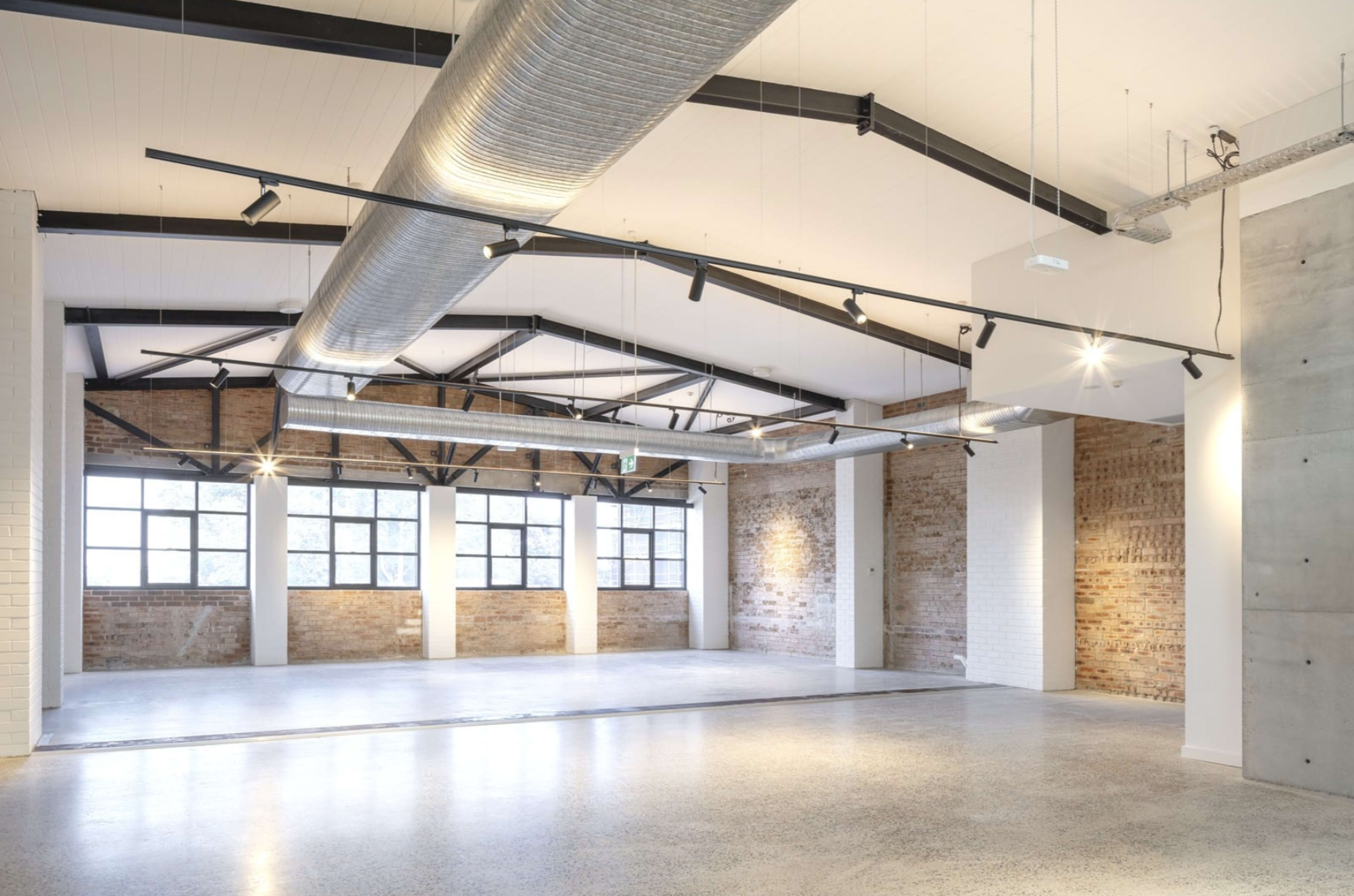 A spacious, industrial-style interior with exposed brick walls, large windows, and a high ceiling featuring visible ductwork and track lighting.