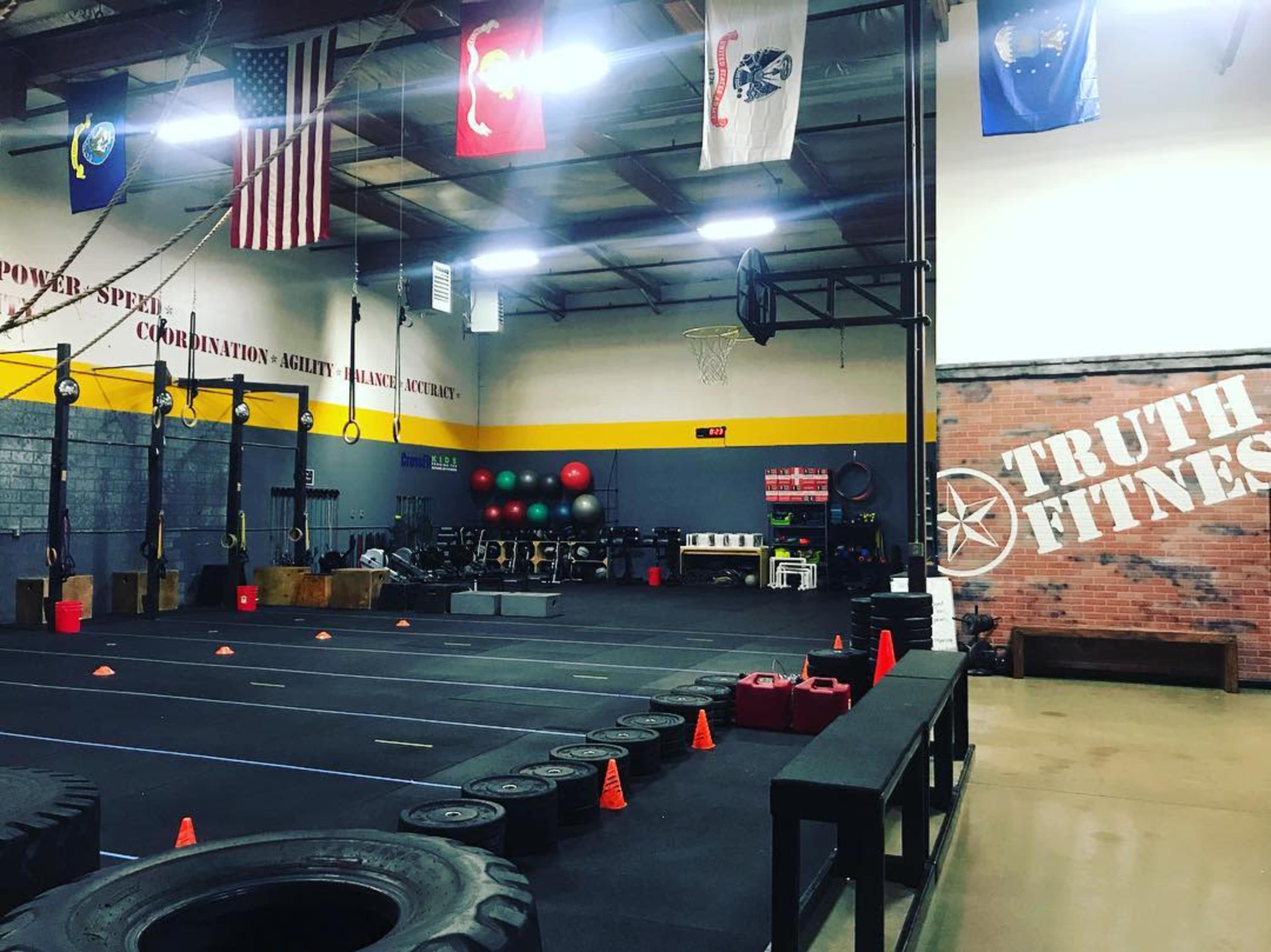 The image shows a spacious indoor gym with exercise equipment, fitness mats, and various colored exercise balls, along with hanging flags and a wall displaying "TRUTH FITNESS."