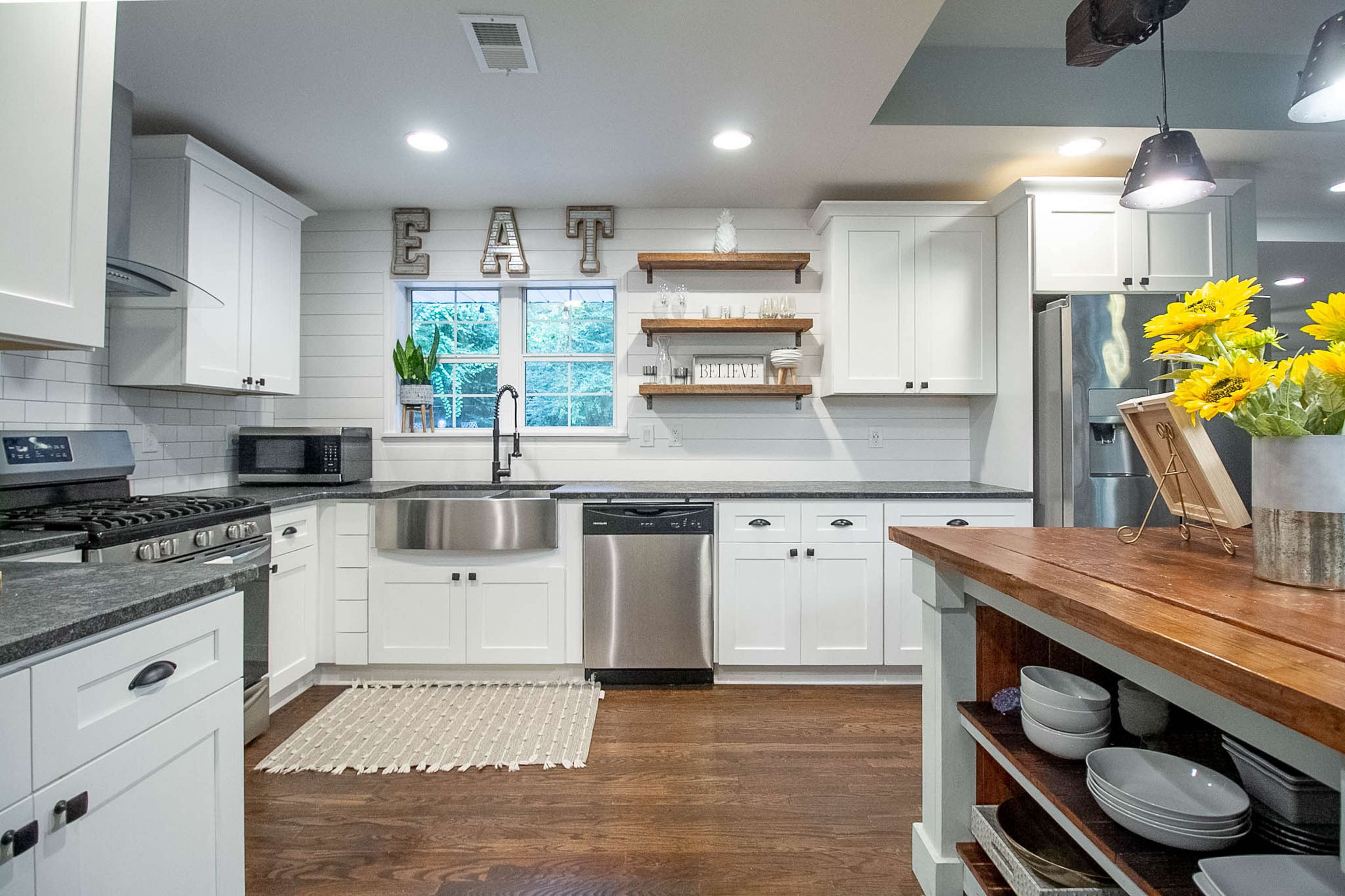 A modern kitchen features white cabinetry, stainless steel appliances, and a wooden table with flowers, centered around a window with decorative shelves.