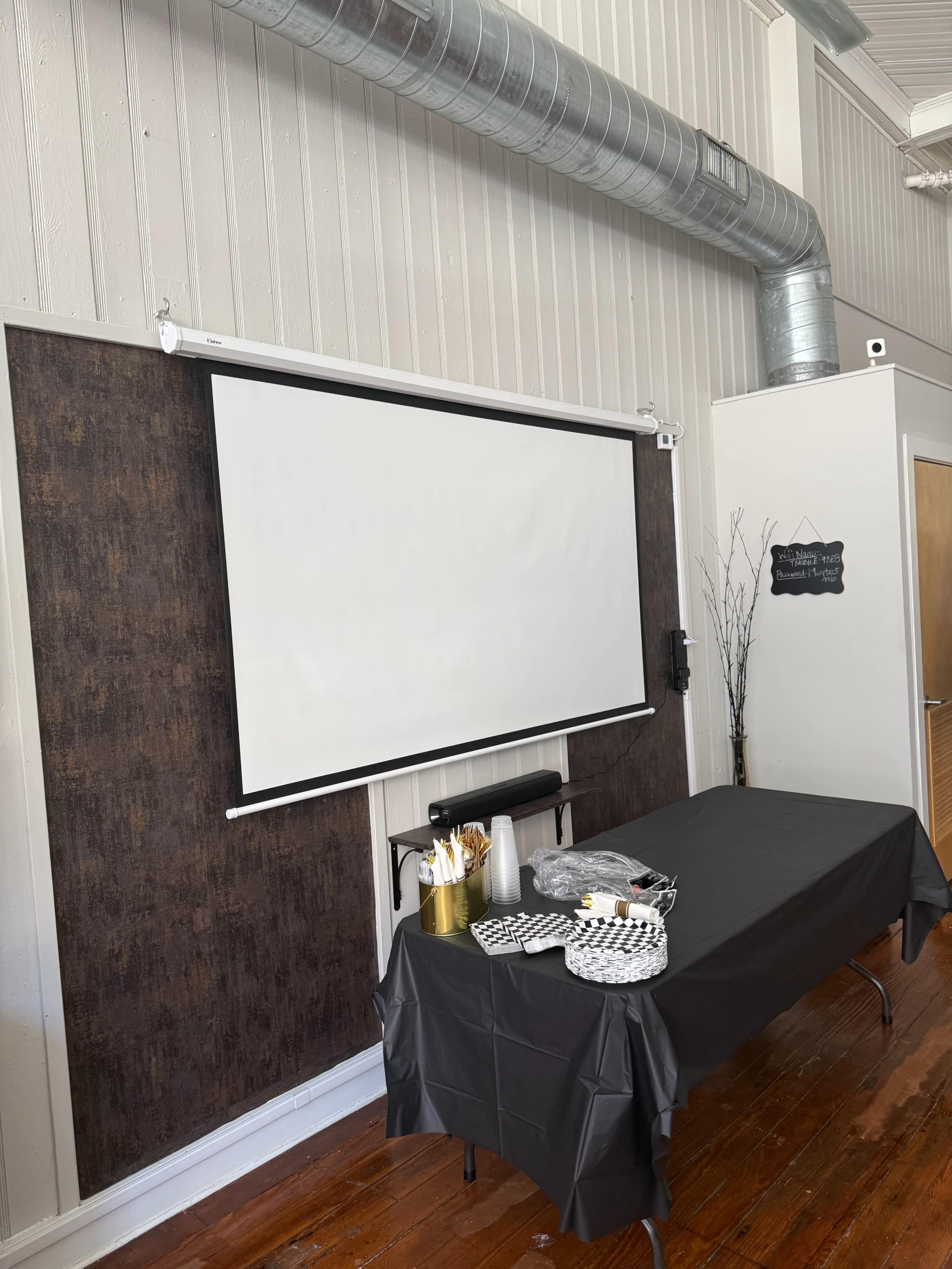The image shows a meeting room with a blank projection screen, a black table covered with a tablecloth, and a selection of utensils and snacks arranged on it.