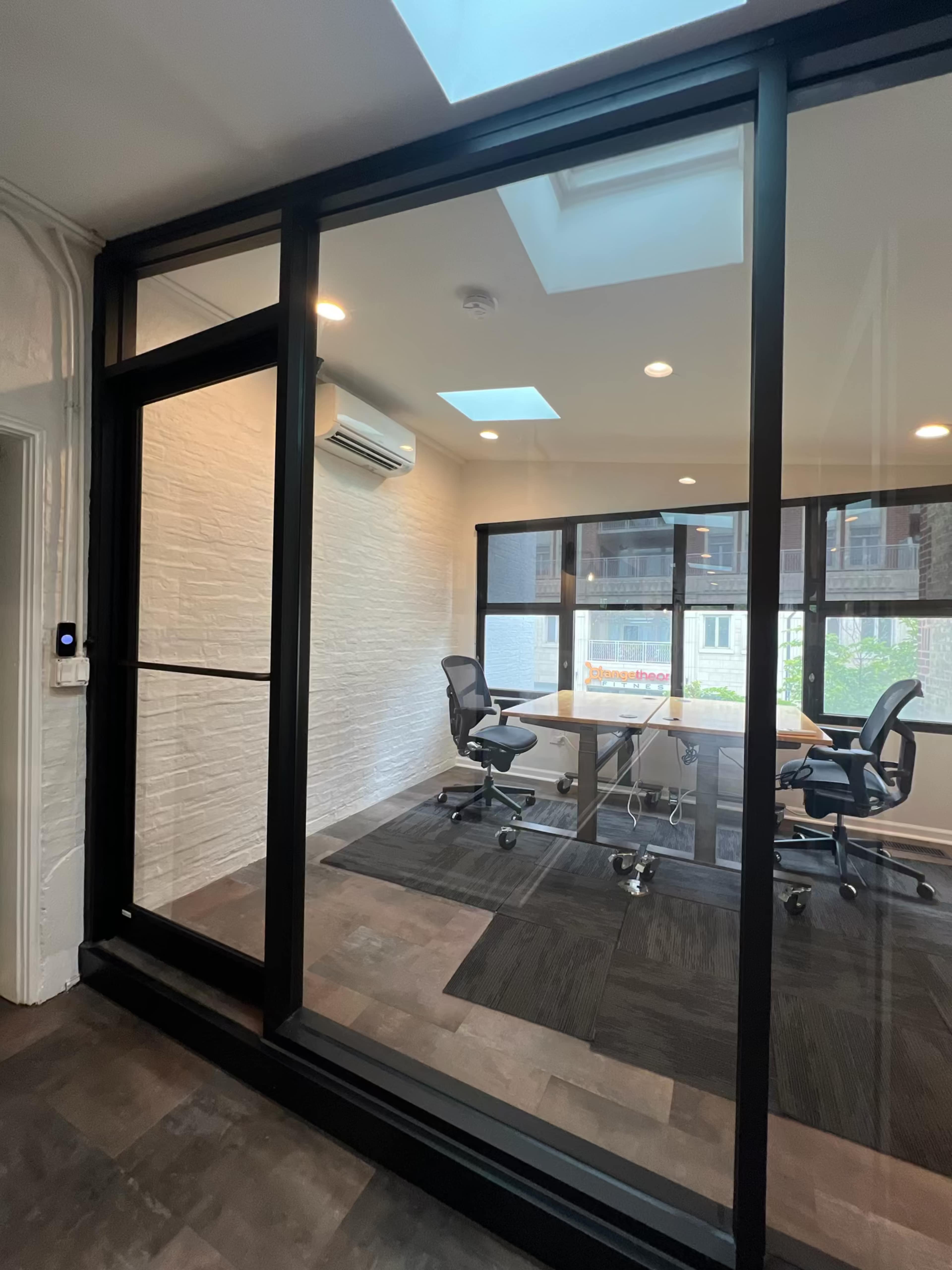 A modern glass-walled conference room with a table and chairs, illuminated by natural light from a skylight.