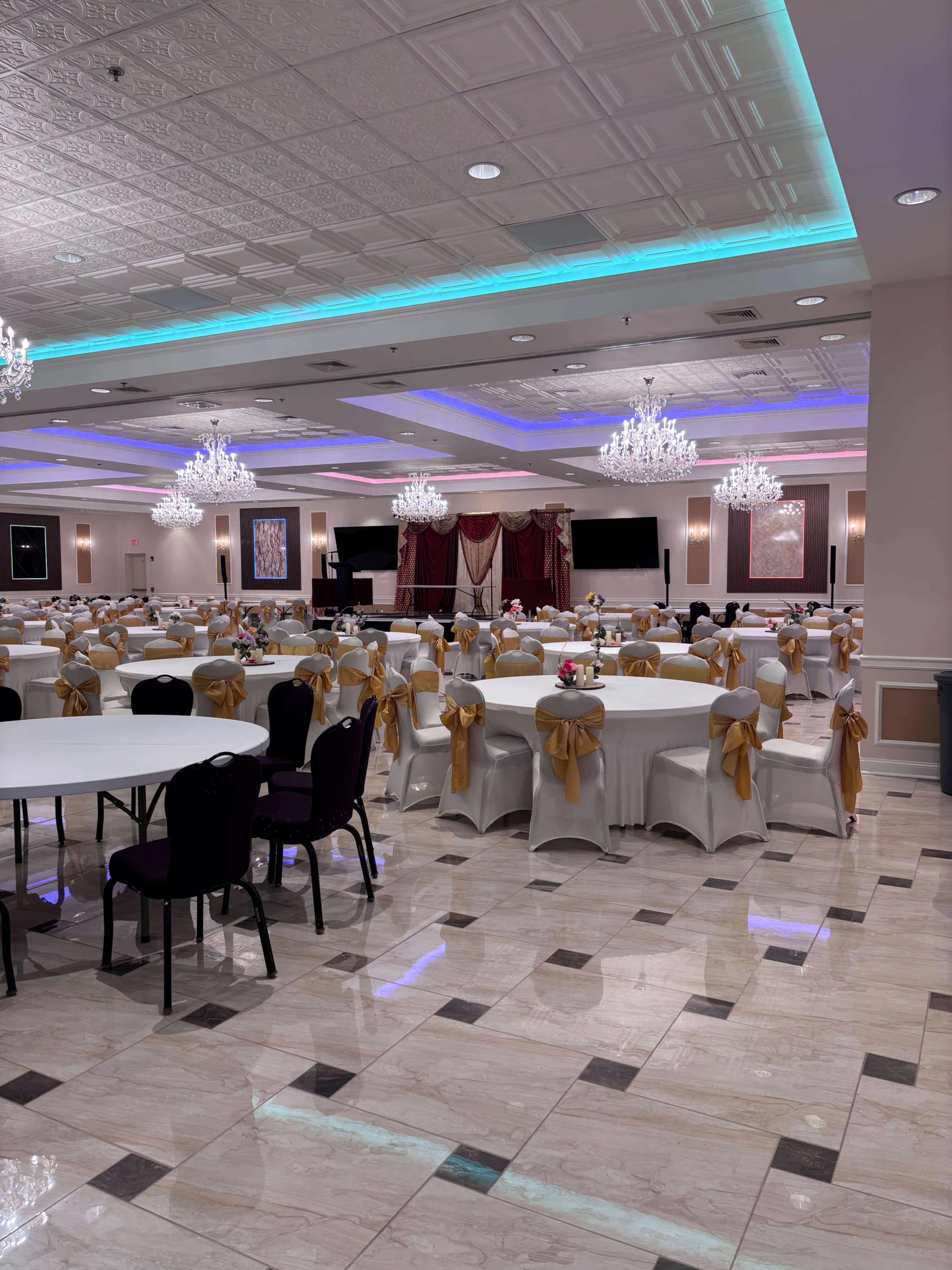 The image shows a spacious banquet hall with round tables adorned with white tablecloths and golden sashes, crystal chandeliers hanging from the ceiling, and a stage area at the back.