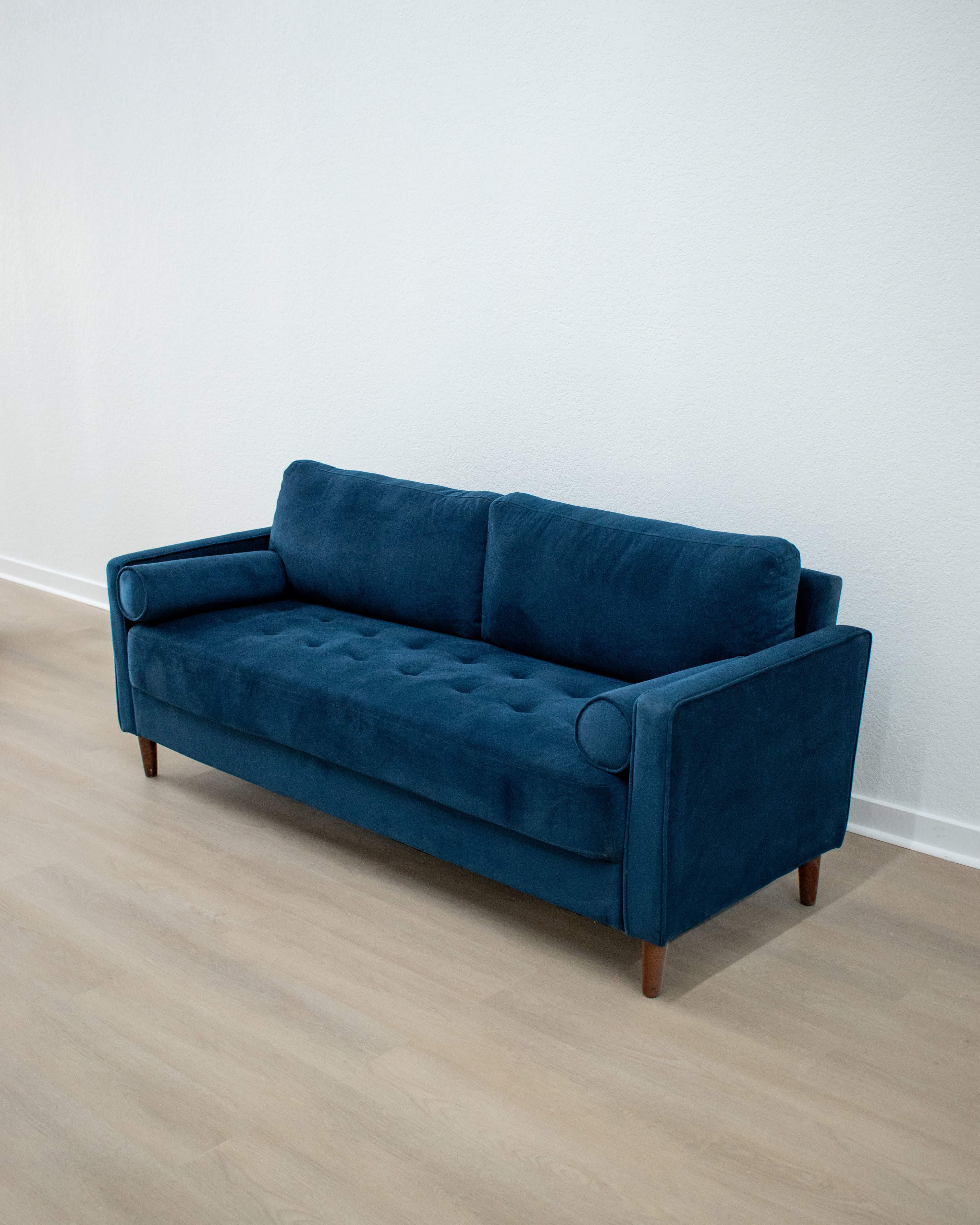 A blue velvet sofa with wooden legs sits against a plain wall on a light wooden floor.