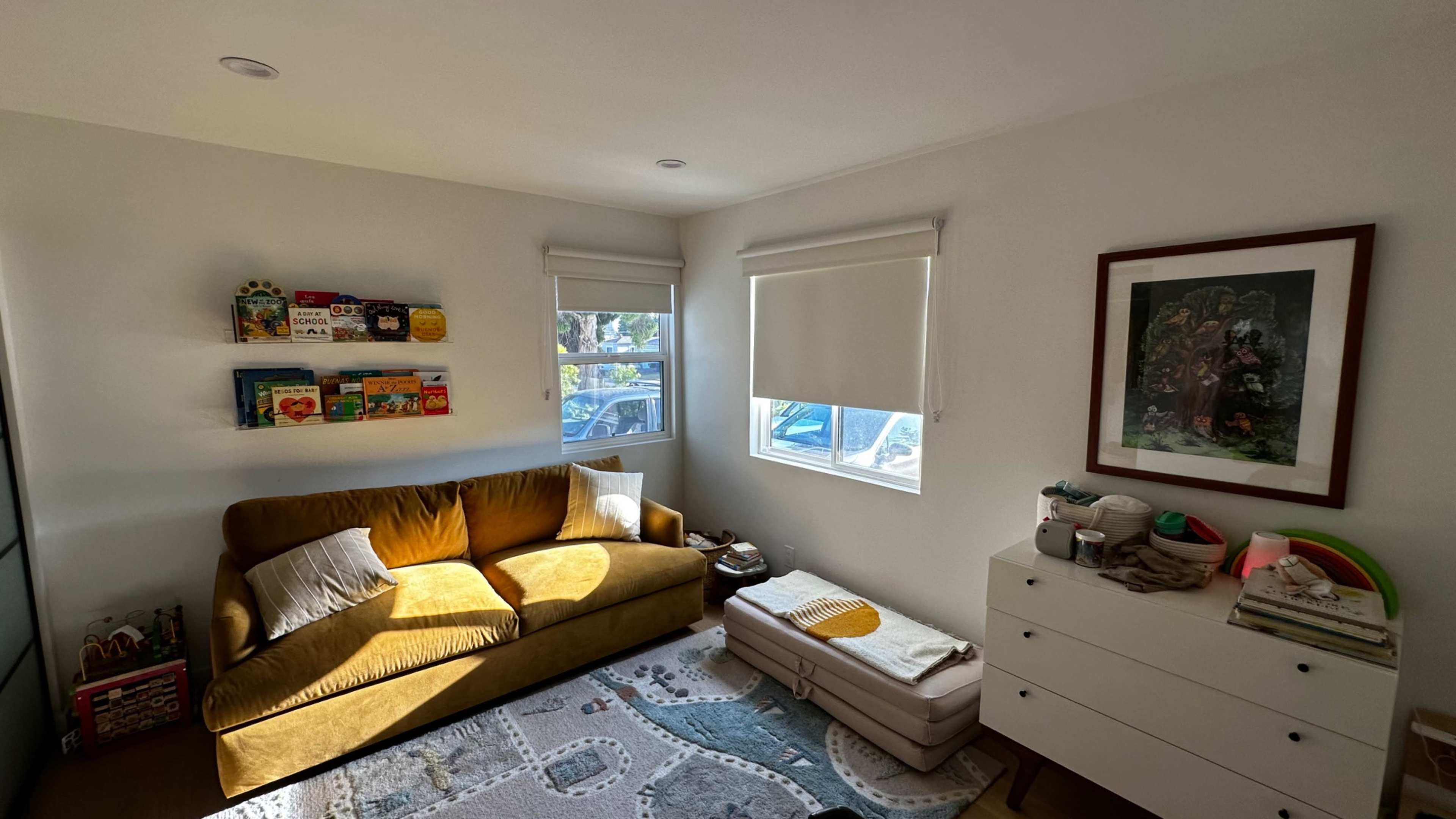 A cozy living room with a yellow sofa, a white dresser, and shelves displaying colorful books on the walls.