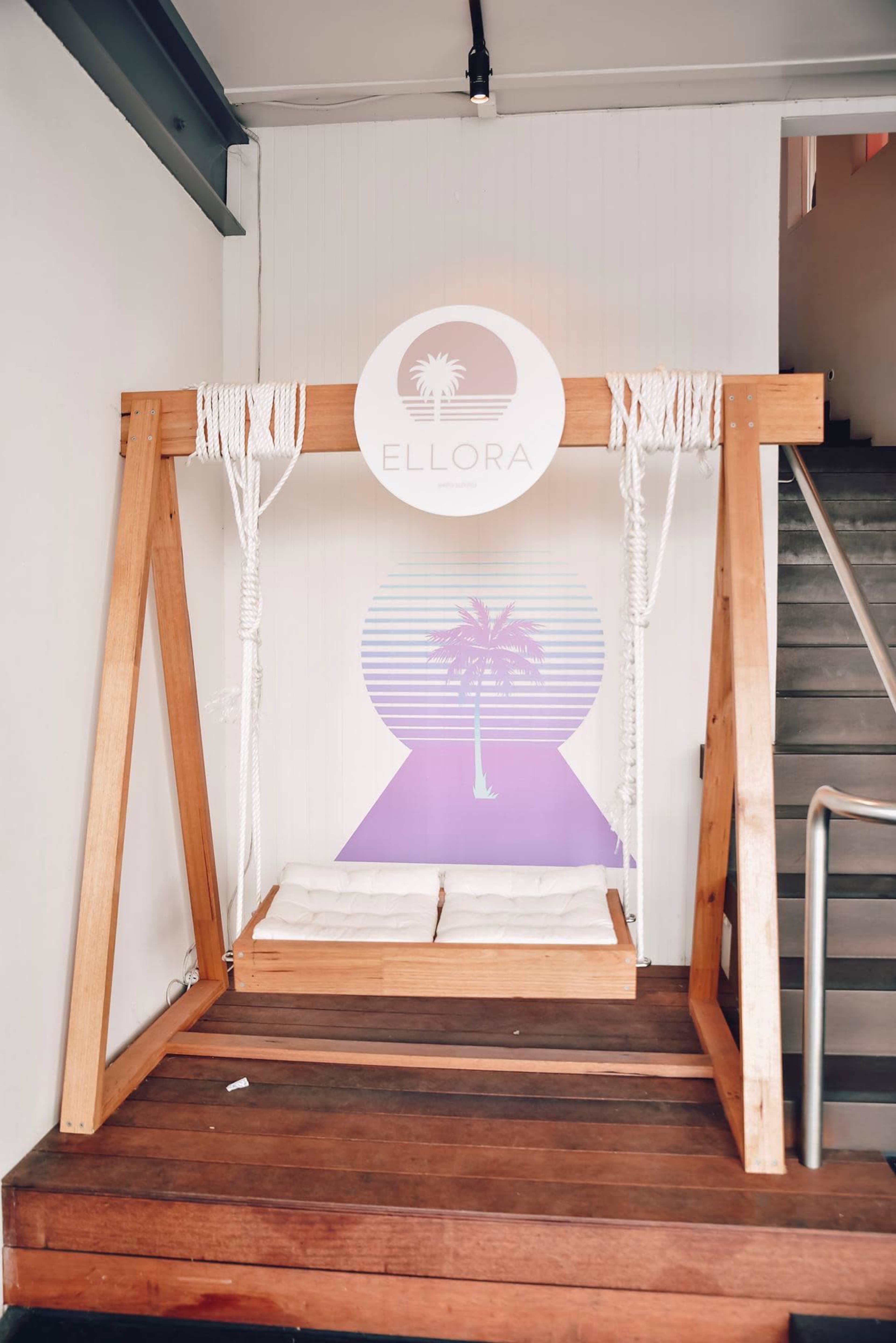 A wooden swing is positioned near a staircase, featuring a circular sign above it that reads "ELLORA" and a mural of a palm tree in the background.
