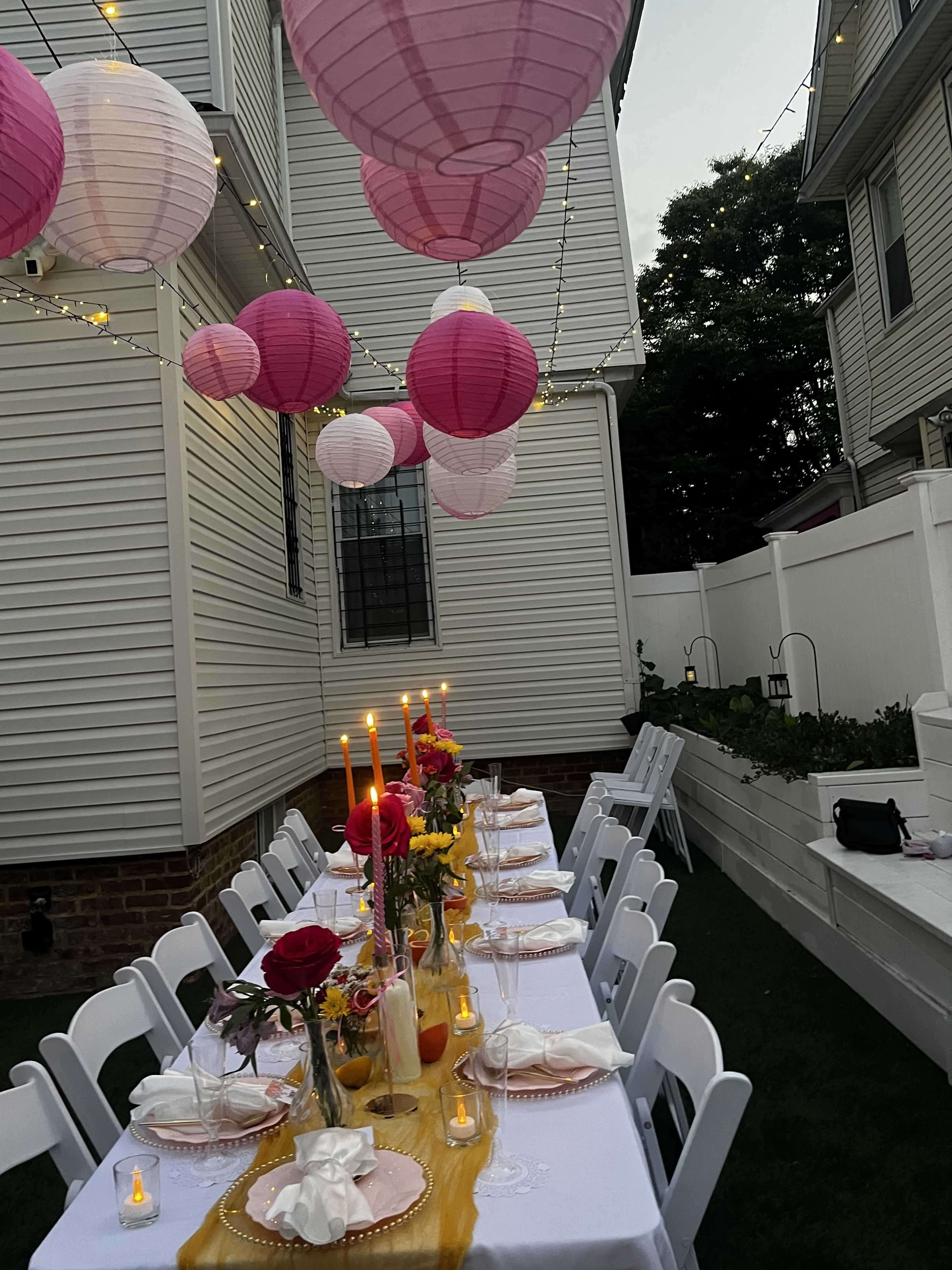 A long dining table is set outdoors with pink lanterns hanging above, surrounded by white chairs and decorated with flowers and candles.