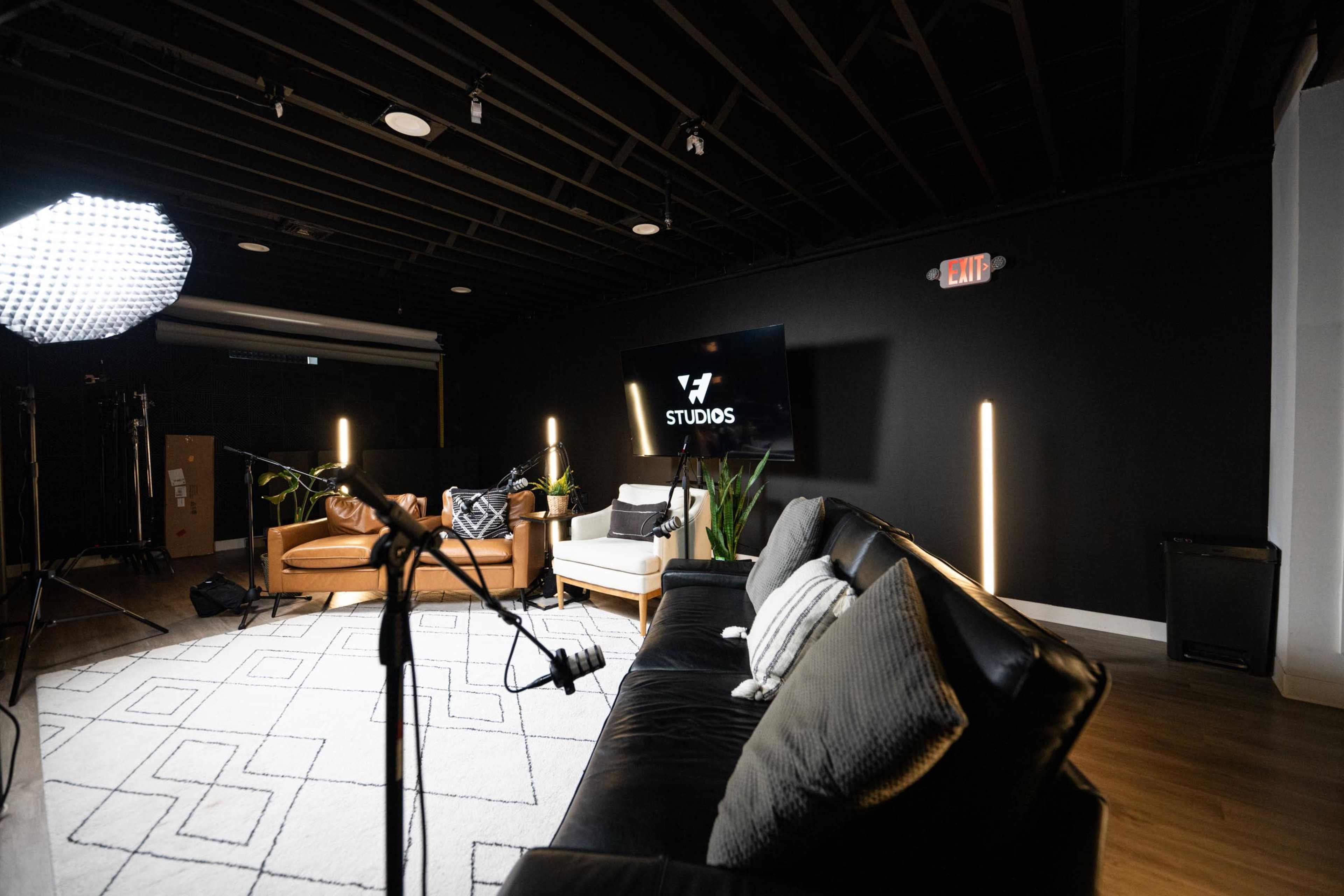The image shows a modern studio setup featuring a black wall, a sofa, two chairs, a large screen displaying the word "STUDIOS," and various lighting equipment.