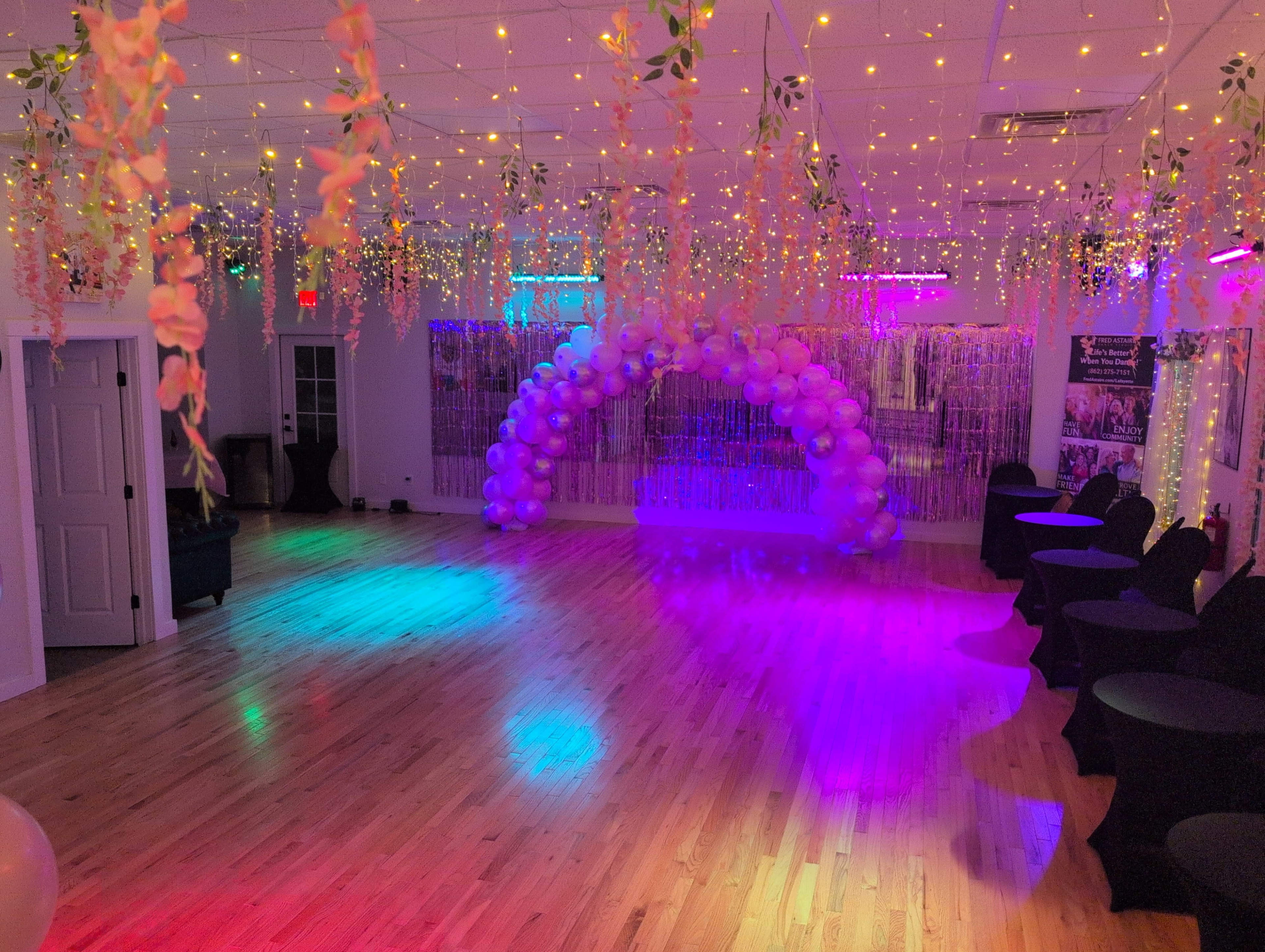The image shows a decorated event space with balloons, string lights, and black tables arranged around a wooden floor.