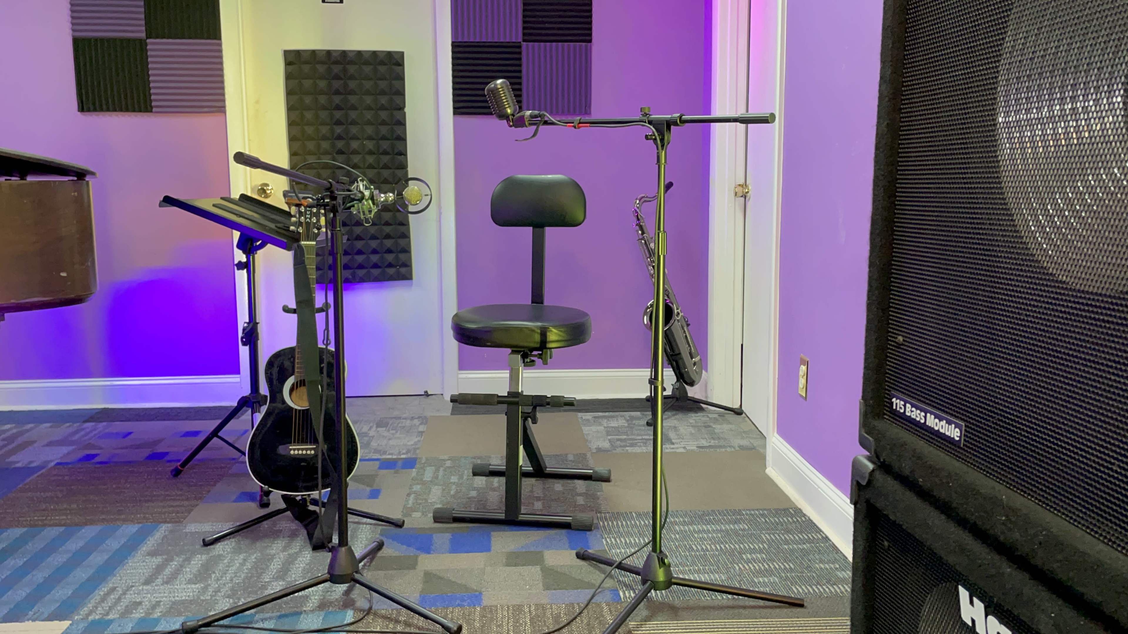 A music rehearsal space featuring a microphone setup, a stool, and several musical instruments, including guitars, against a purple wall.