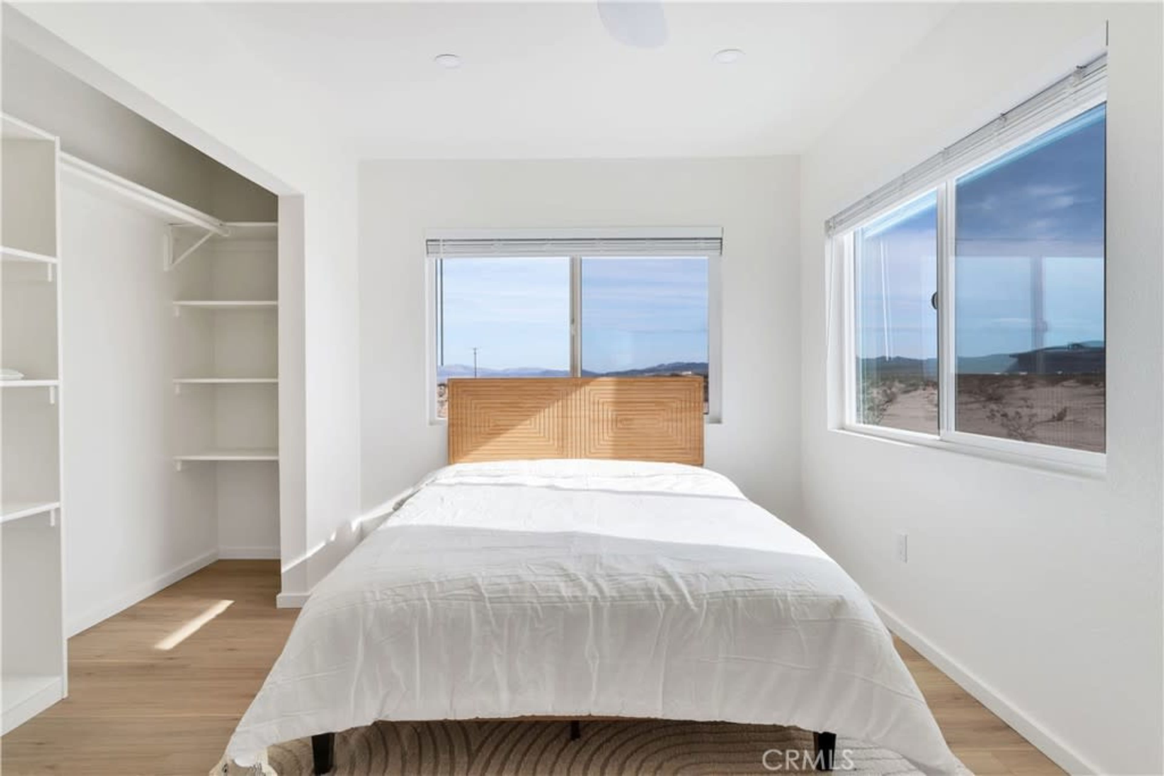 A bright bedroom featuring a bed with a wooden headboard, white bedding, and a large window overlooking a desert landscape.