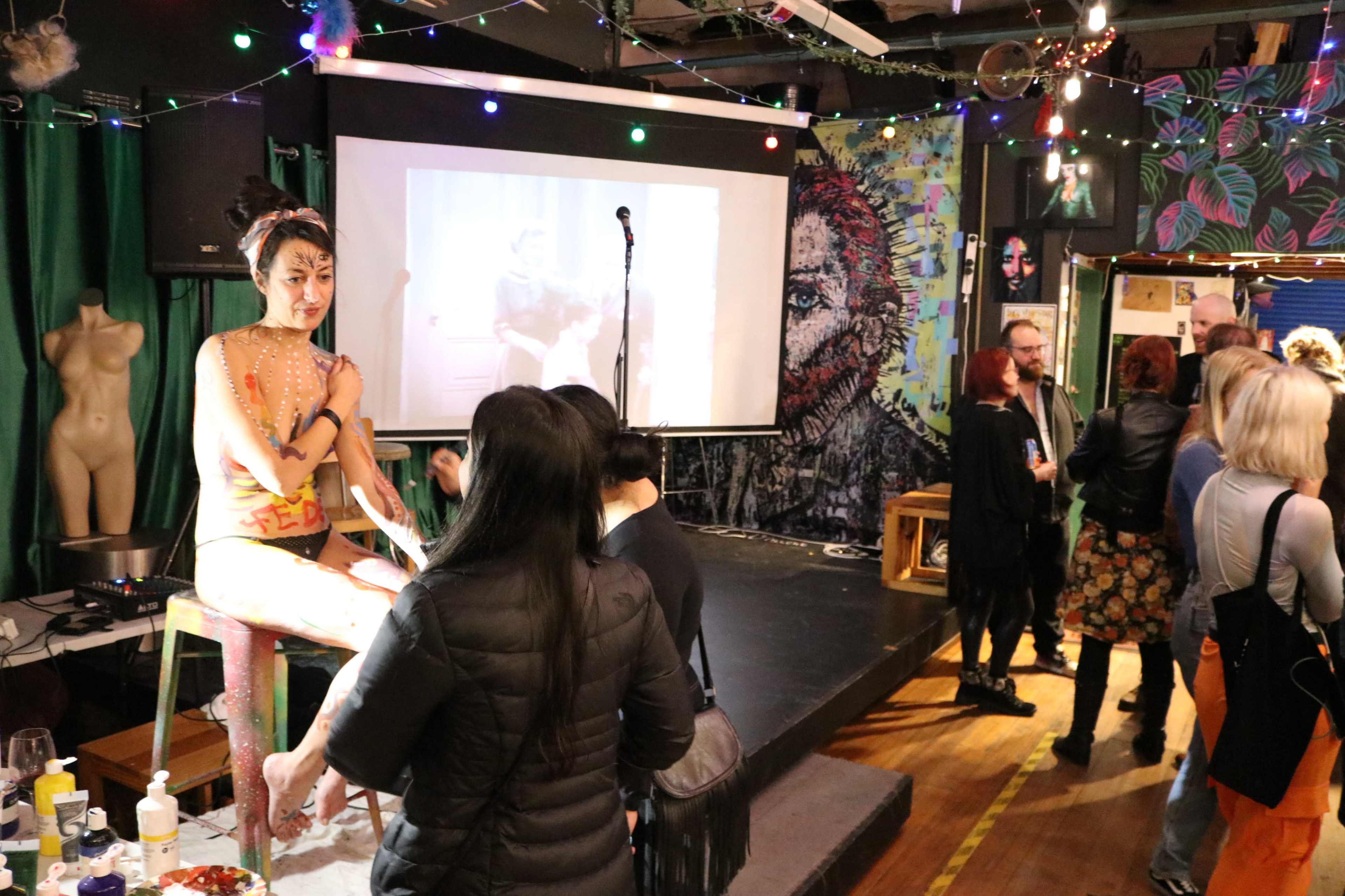 A woman with body paint sits on a stool while people engage with each other and a video plays on a screen in a colorful venue.