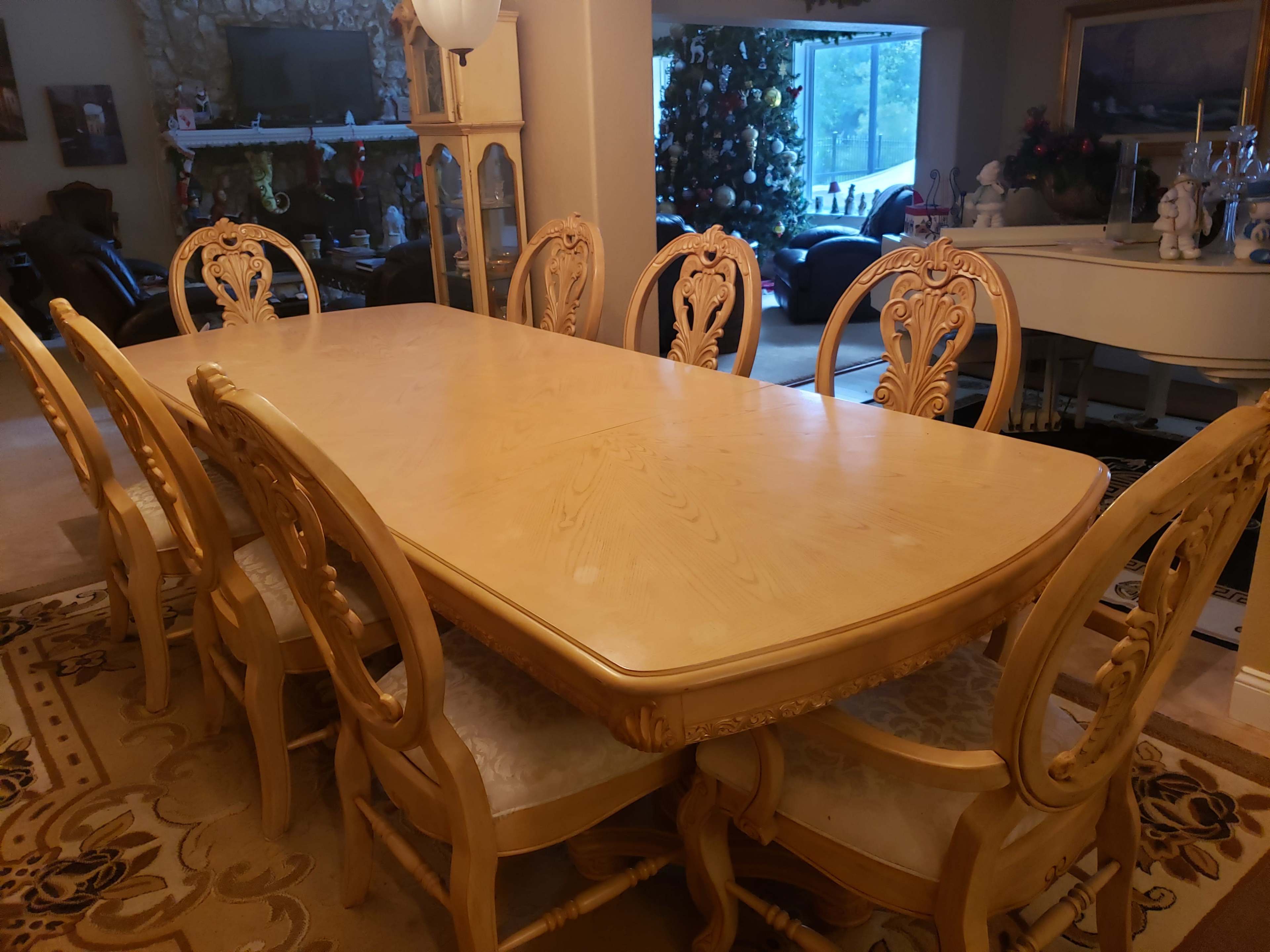 A wooden dining table is surrounded by six intricately designed chairs in a well-lit room with a decorated Christmas tree visible in the background.