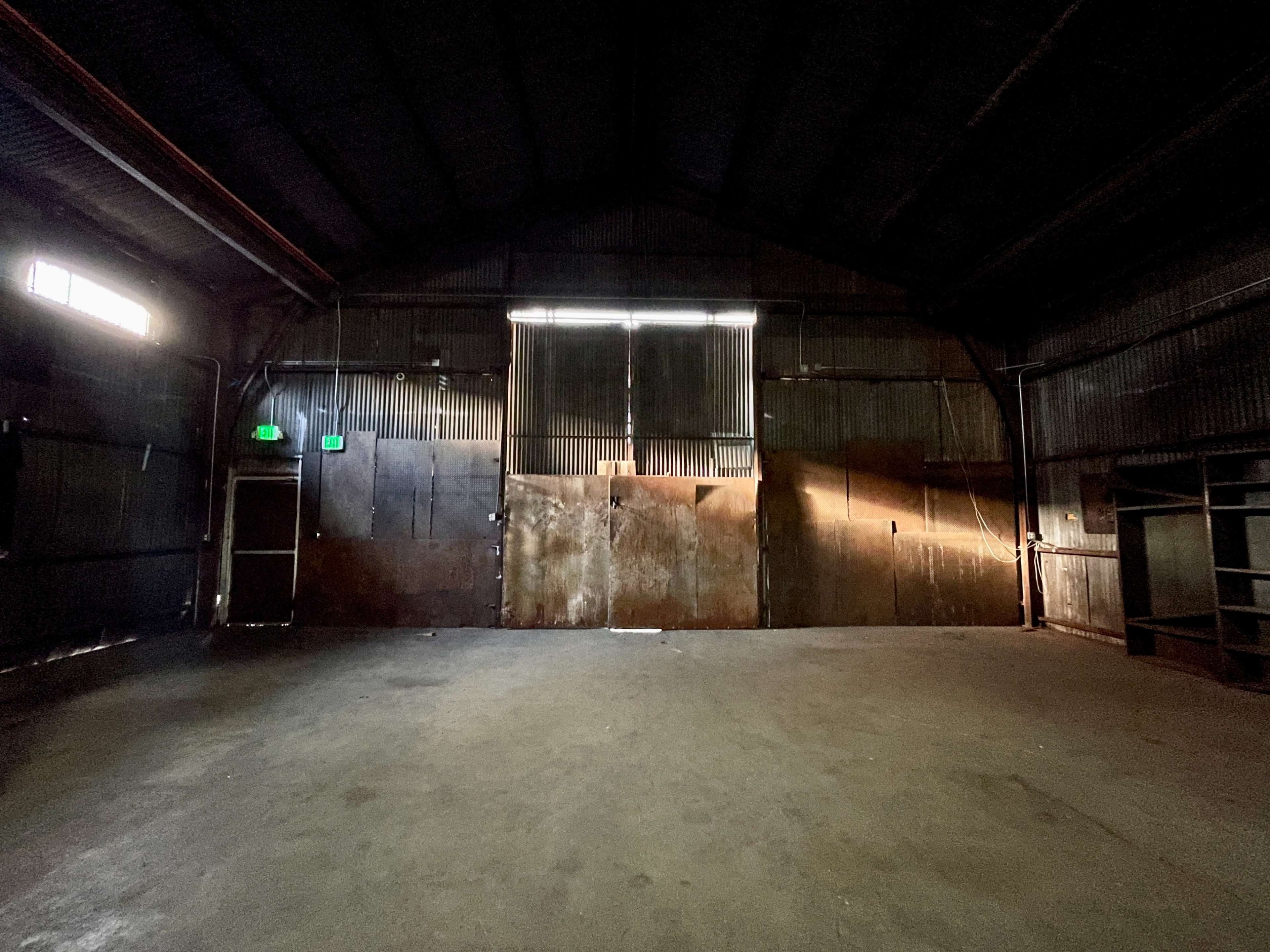 The interior of a large, empty warehouse with metal walls and dim light filtering through partially open doors.