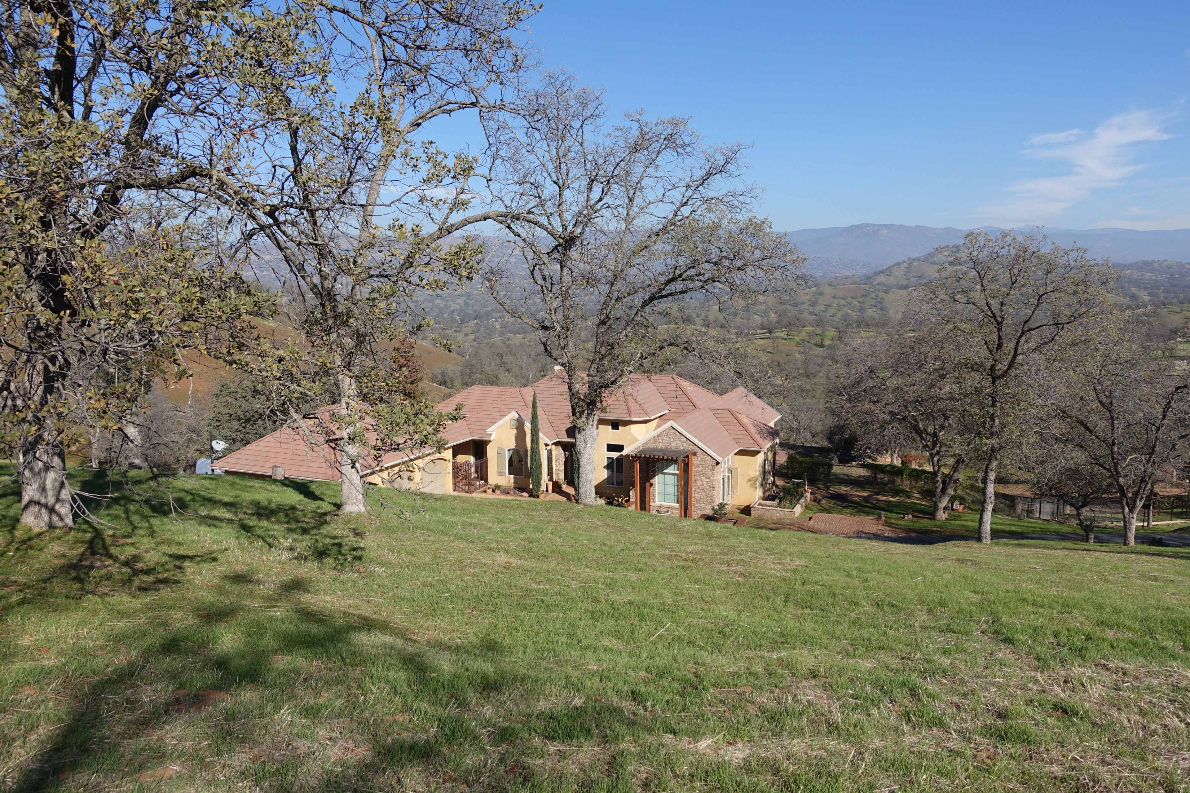 A large, single-story house with a tiled roof is situated on a grassy hillside, surrounded by trees and overlooking a valley.