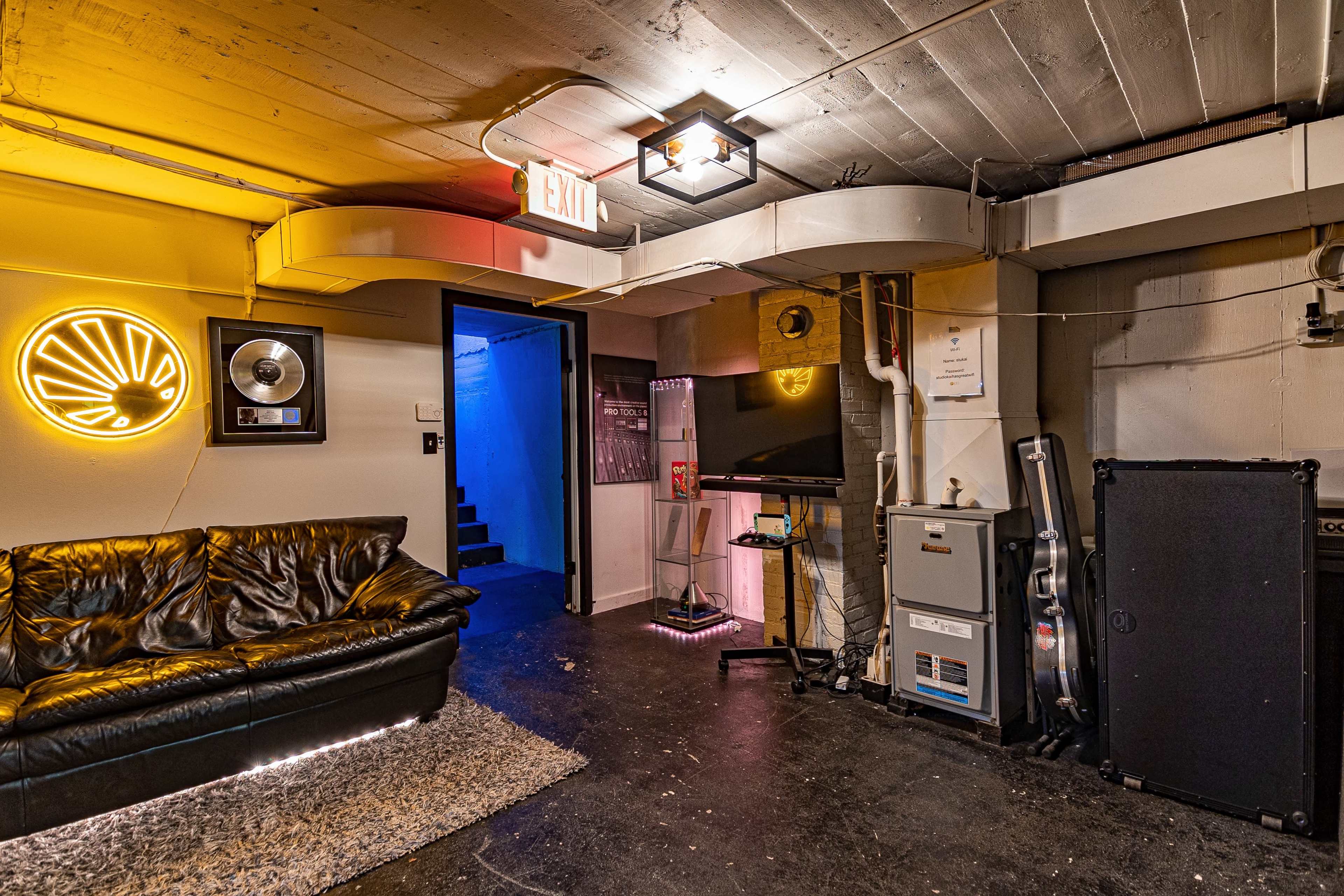 A basement room with a black leather couch, a neon wall decoration, and a small television beside a staircase leading up.