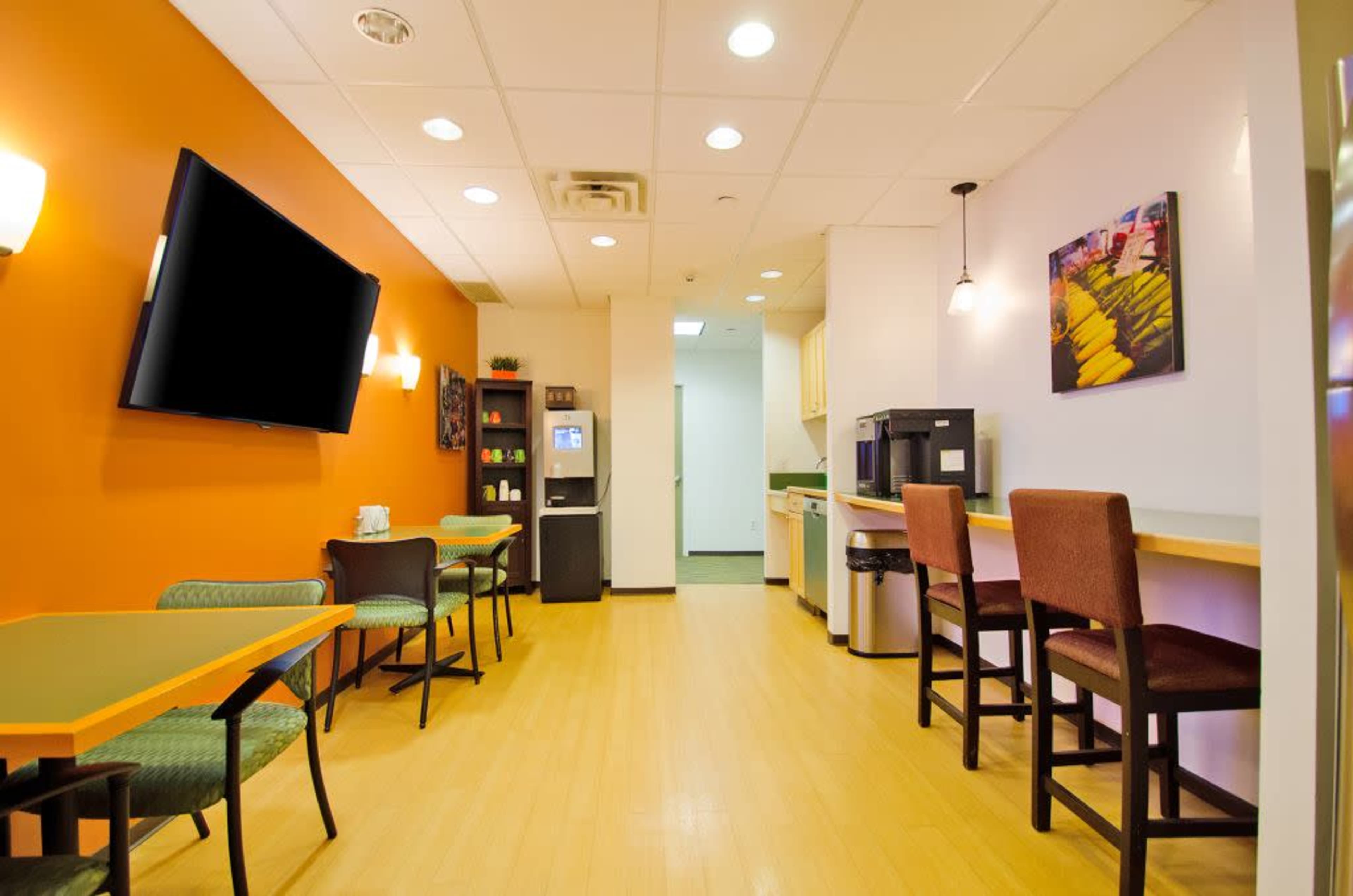 A modern break room with orange walls, a television mounted on one side, a refrigerator, a coffee machine, and seating areas with tables and chairs.