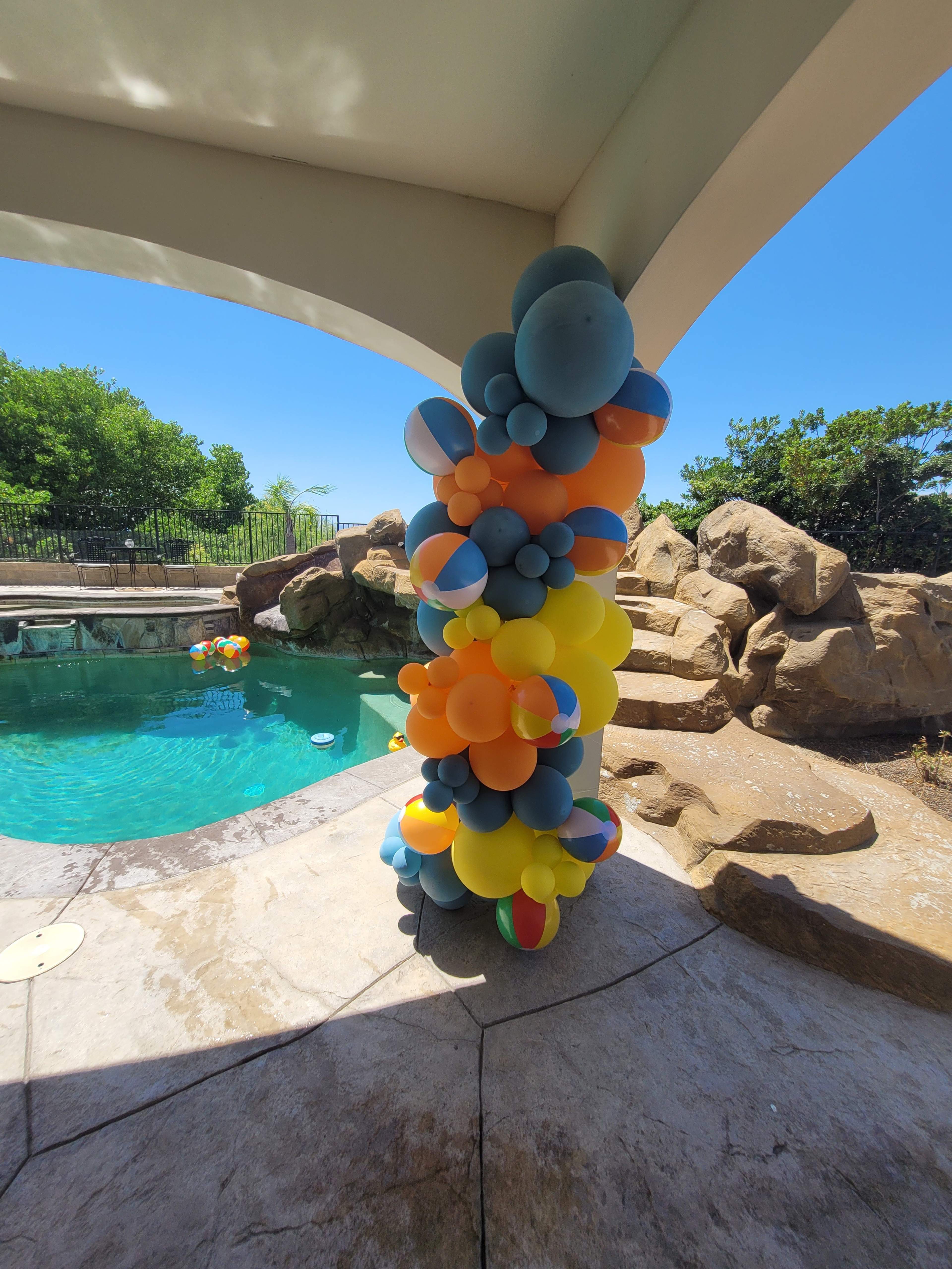 A column of colorful balloons stands next to a swimming pool surrounded by rocks and greenery under a clear blue sky.