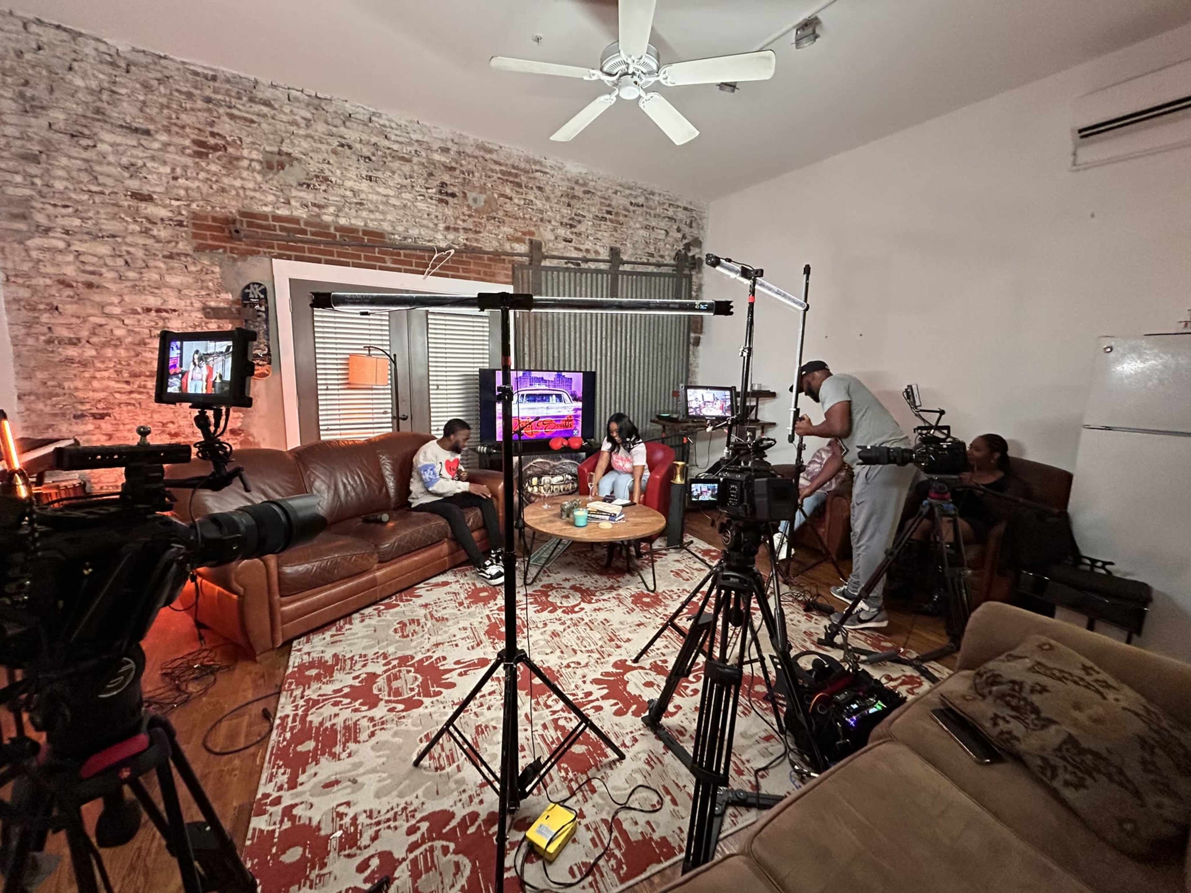 A recording studio setup with multiple cameras focused on two people seated on a couch, surrounded by lighting equipment and audio devices.