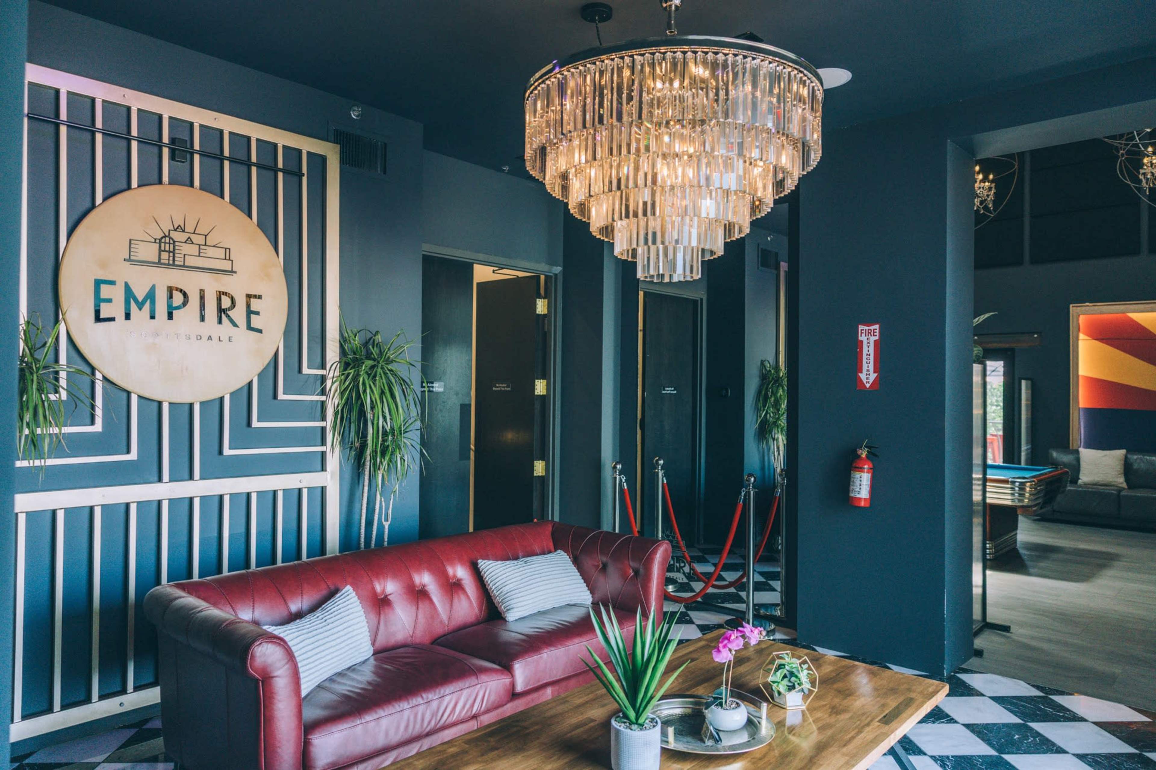 The image shows a modern lobby with a red leather sofa, a large chandelier, and decorative elements like plants and a sign that reads "EMPIRE" on the wall.