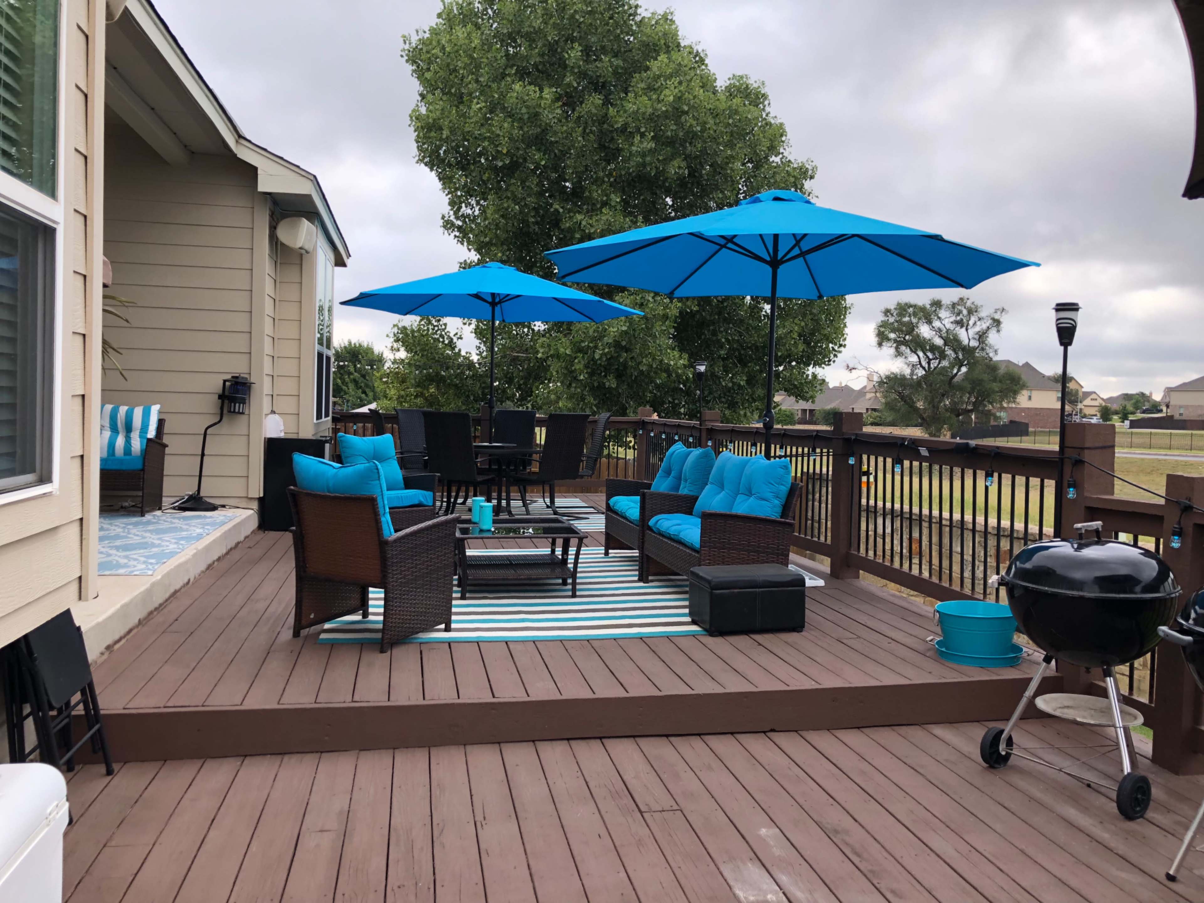 A backyard deck features blue umbrellas over a seating area with blue cushions, a table, and a grill, surrounded by a grassy landscape.