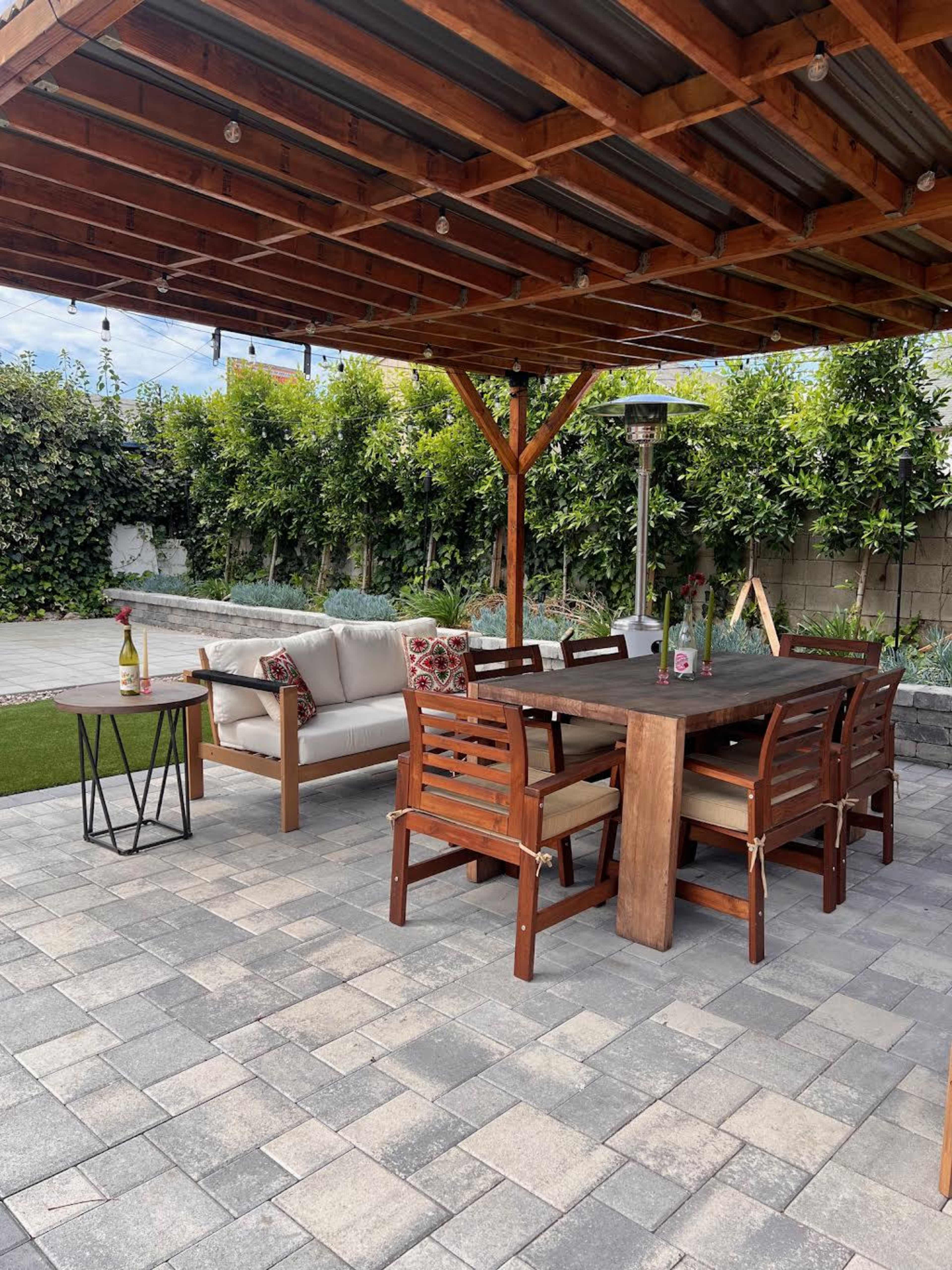 The image shows a patio area featuring a wooden roof, a seating area with a couch and a dining table surrounded by chairs, all on a stone paver surface.