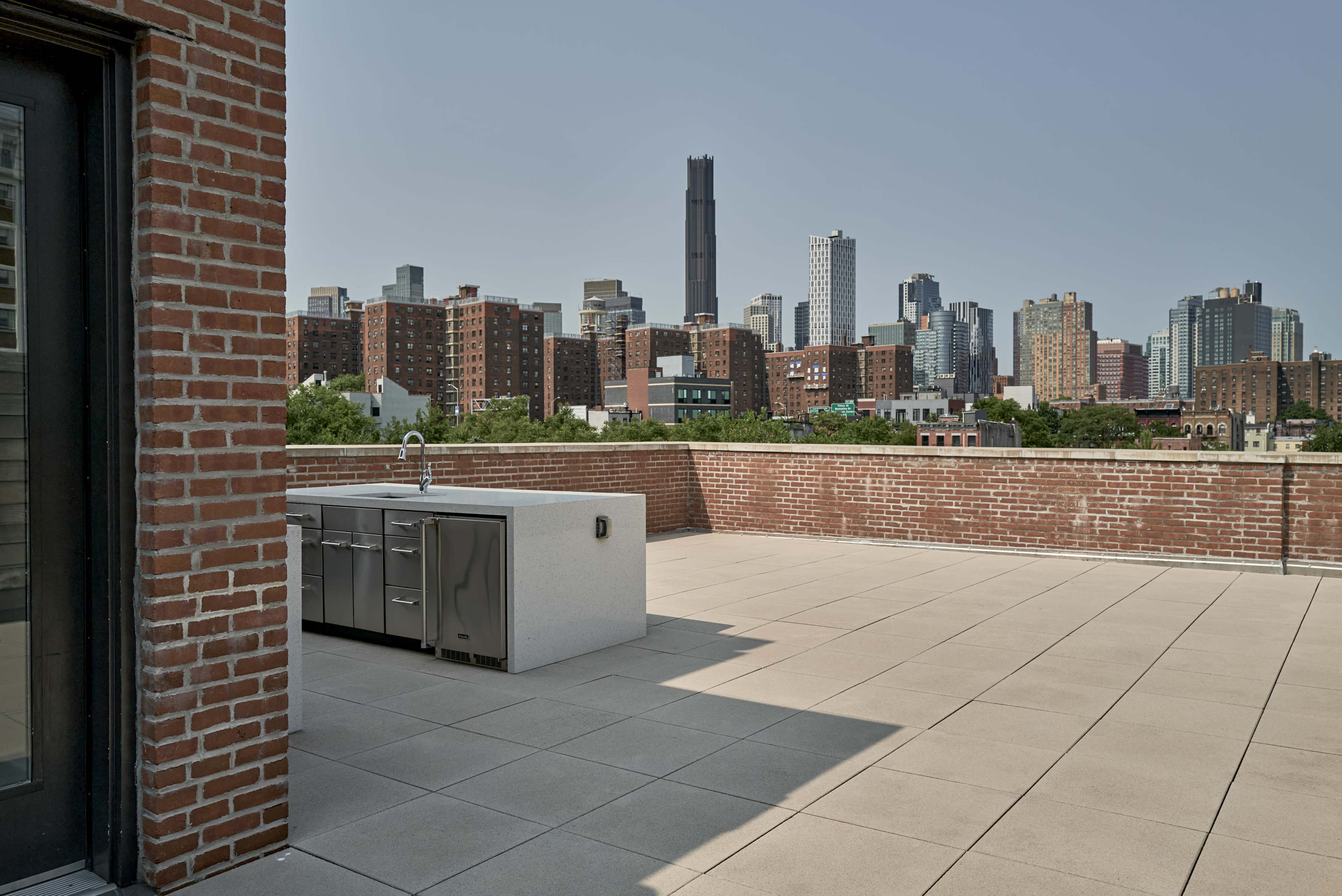 The image shows a rooftop terrace with a modern outdoor kitchen and a skyline view of tall buildings in an urban setting.