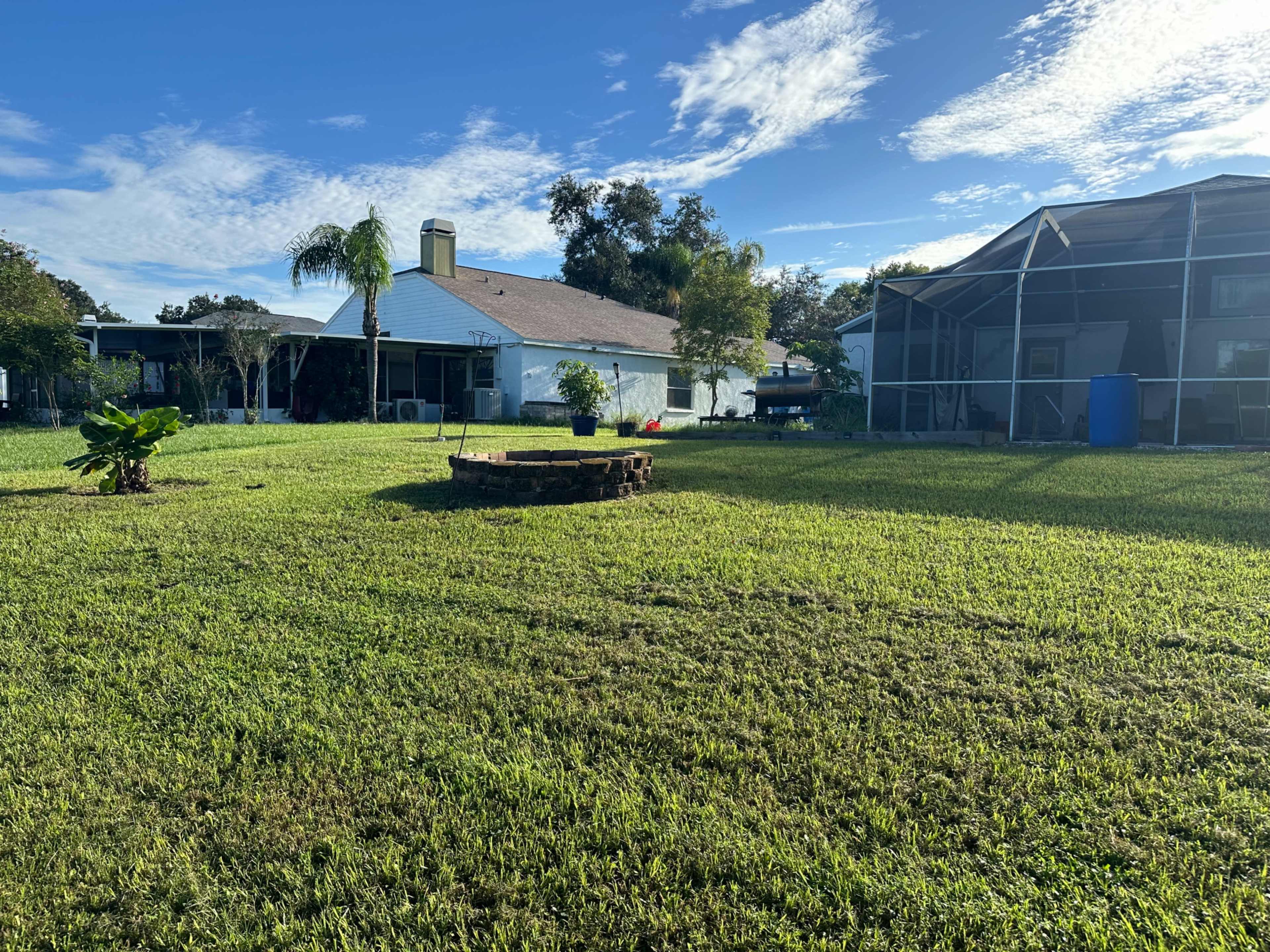 A spacious backyard features a circular stone fire pit surrounded by well-maintained grass, with a house and pool enclosure in the background.