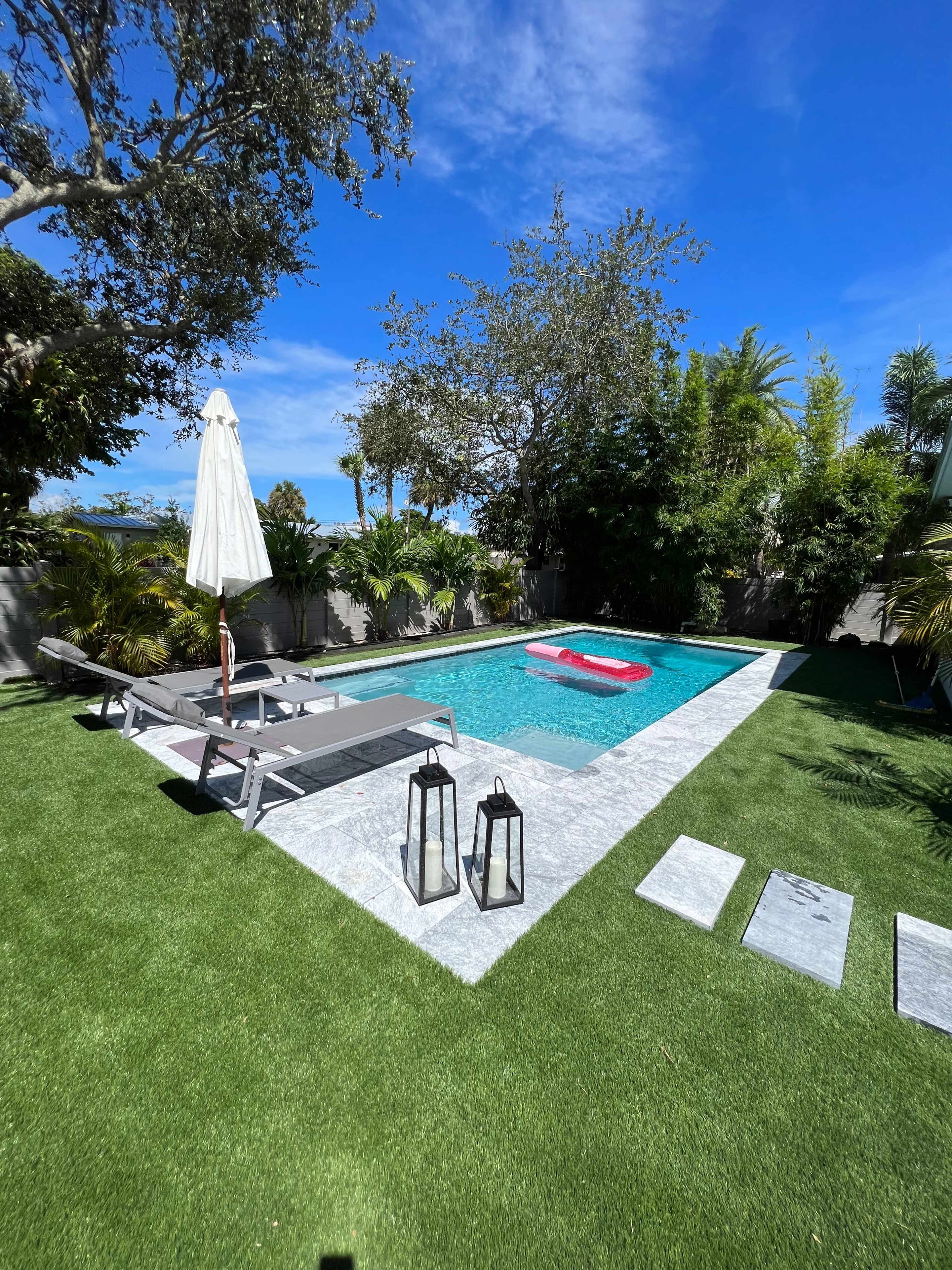 A swimming pool with a lounge area, surrounded by greenery, includes a white umbrella, lounge chairs, and lanterns on a stone path.