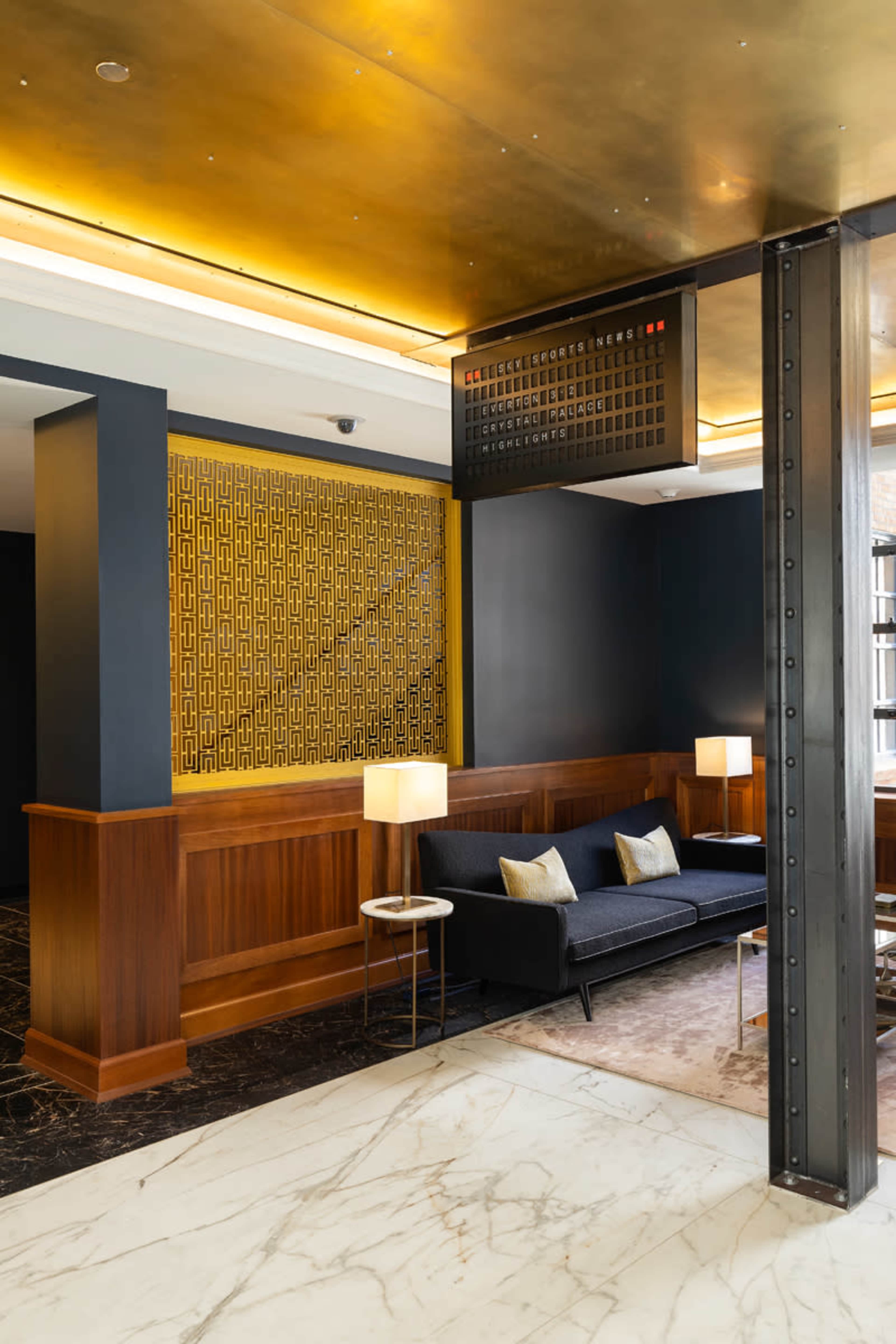 A modern hotel lobby featuring a dark seating area, a wooden reception desk with a decorative screen, and geometric lighting fixtures.