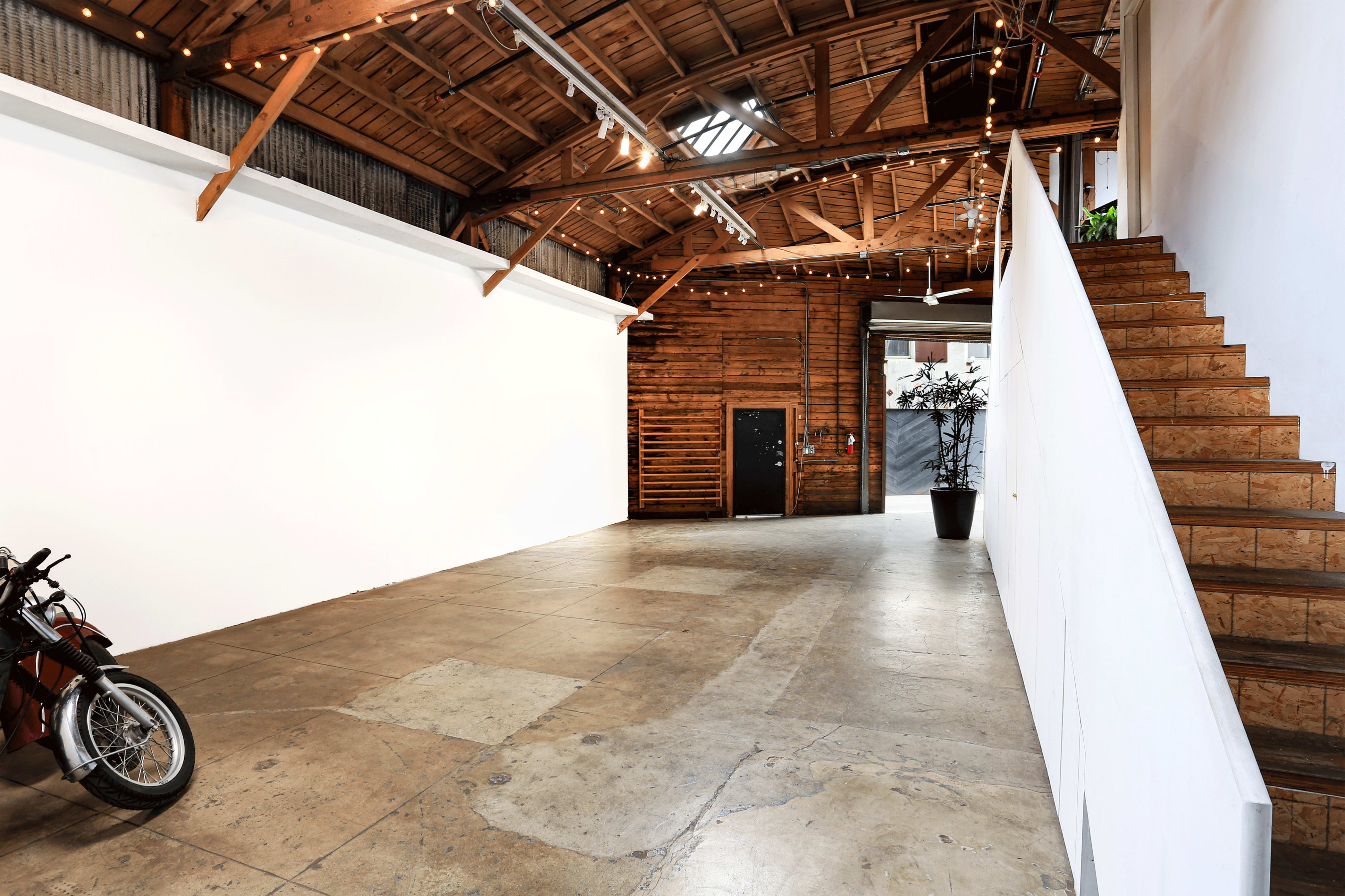 The image shows a spacious interior with a wooden ceiling, a motorcycle parked on the left, and a staircase leading to an upper level on the right.