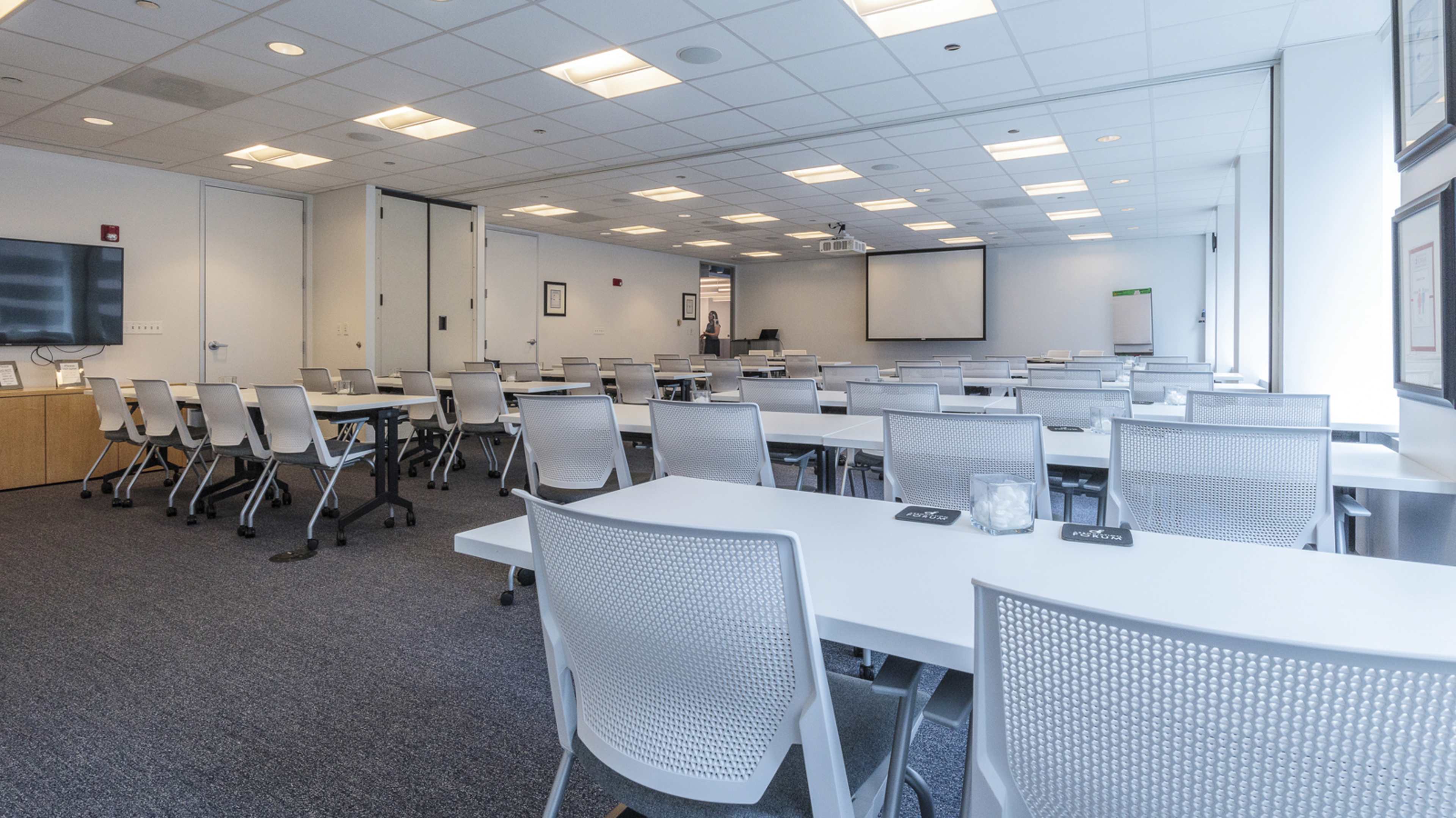 A spacious conference room features rows of white tables and chairs, with a large screen at the front and windows along one wall.