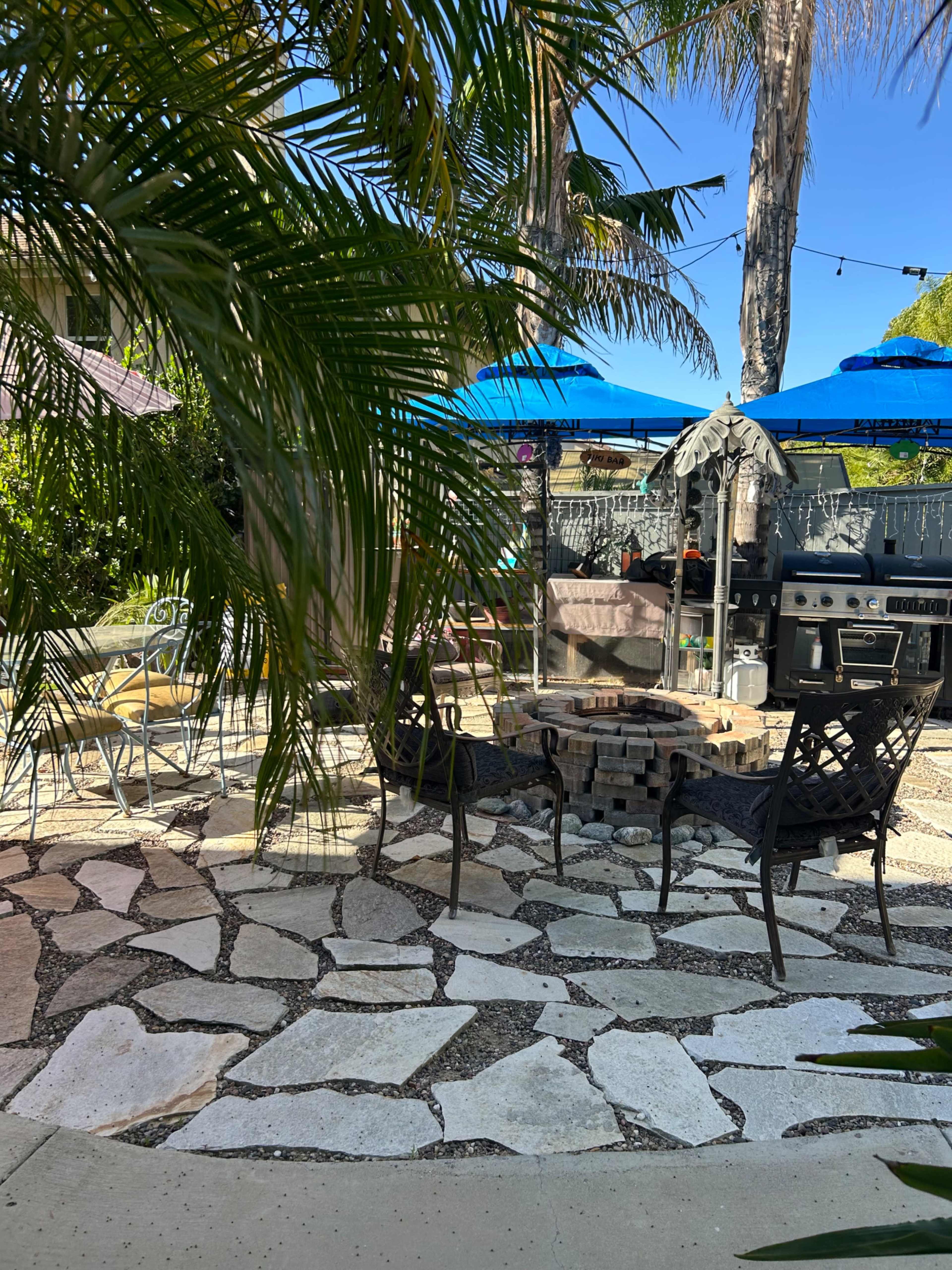 The image shows a backyard area with a stone patio, several outdoor chairs, and a grill under blue umbrellas surrounded by palm trees.