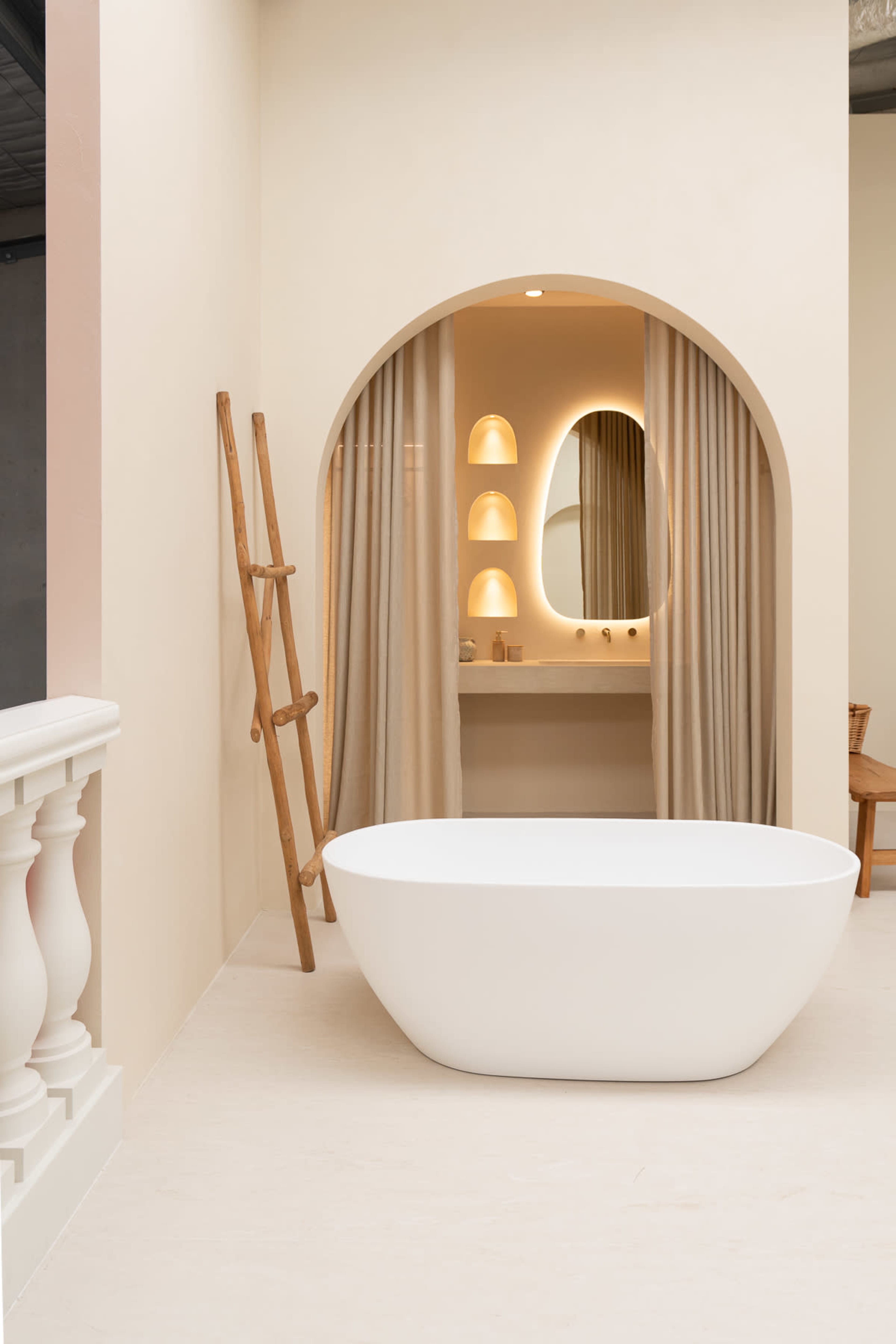 A modern bathroom features a freestanding white bathtub, an arched mirror, and soft lighting, framed by beige walls and curtains.
