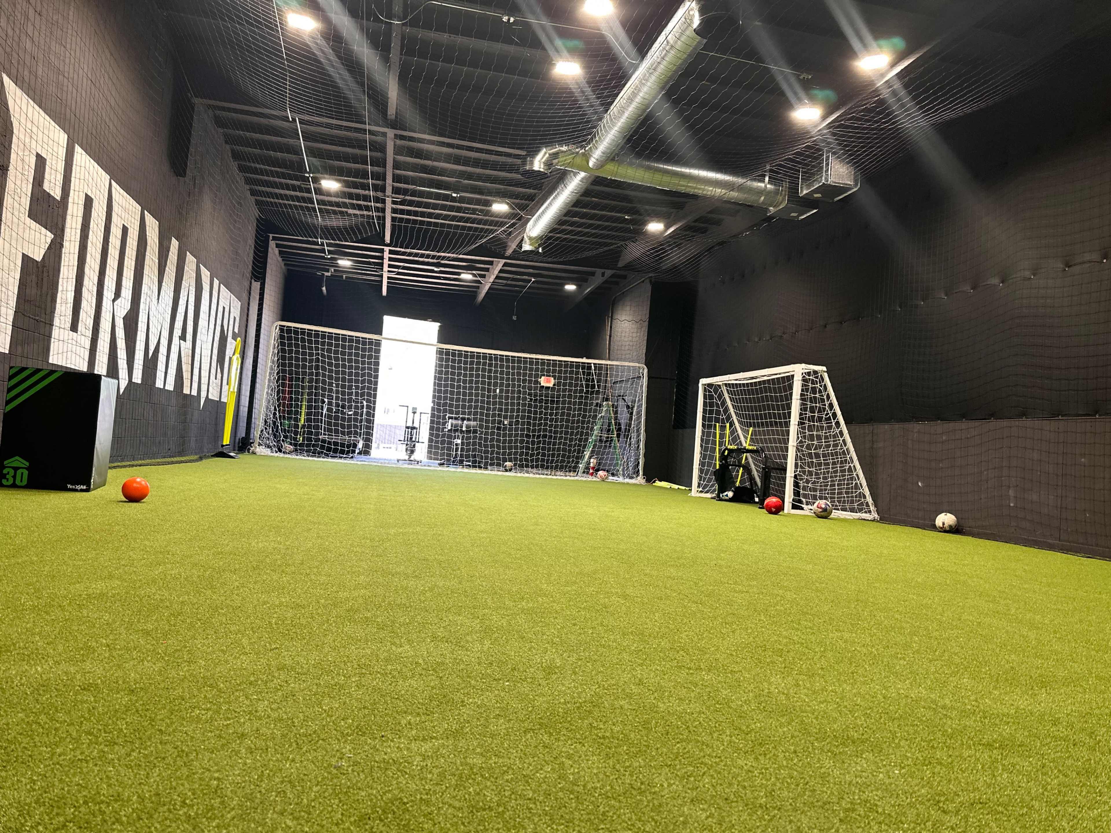 An indoor soccer training facility with artificial turf, two goals, and various sports equipment scattered around.