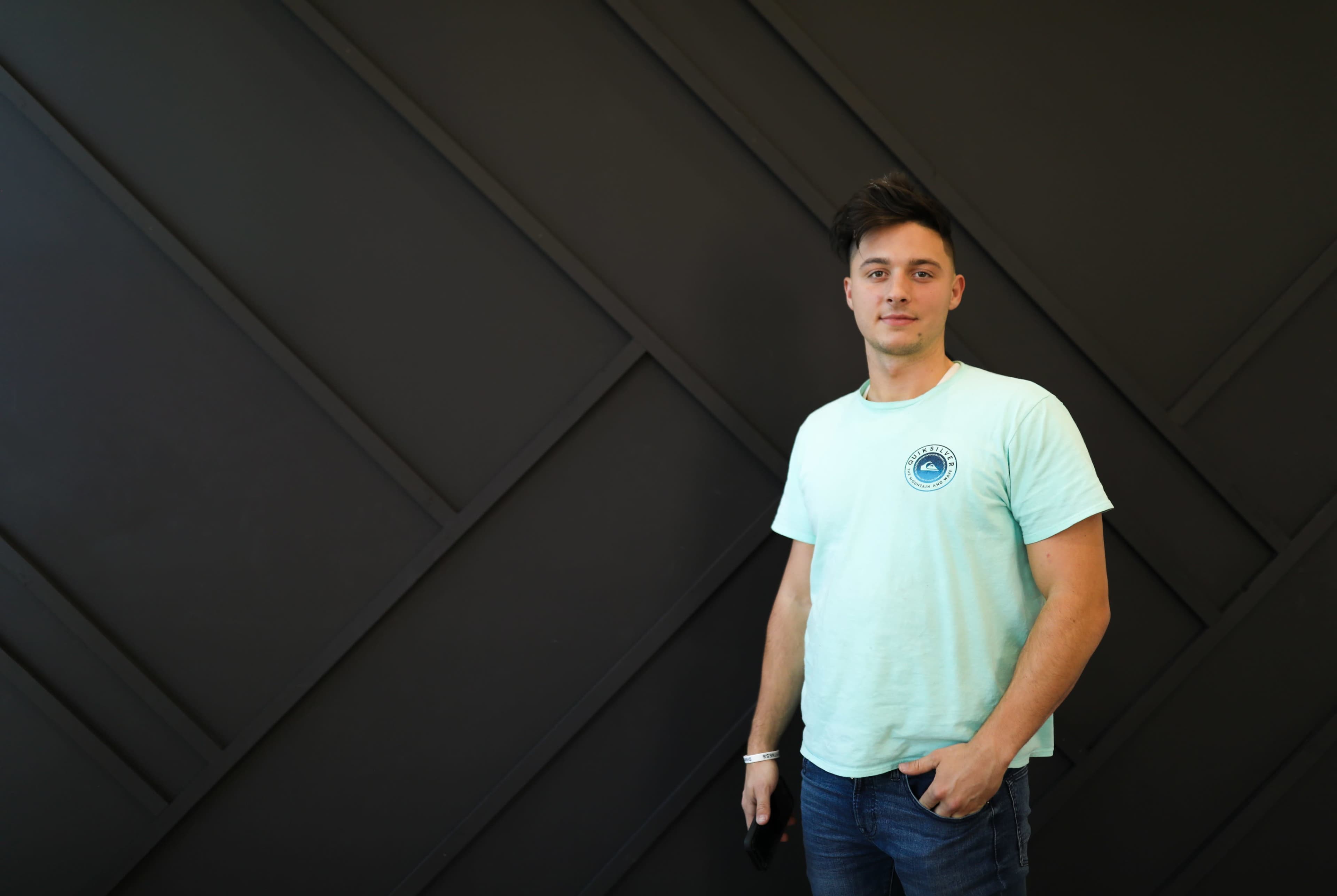 A young man stands against a dark geometric wall while wearing a light blue t-shirt and jeans.