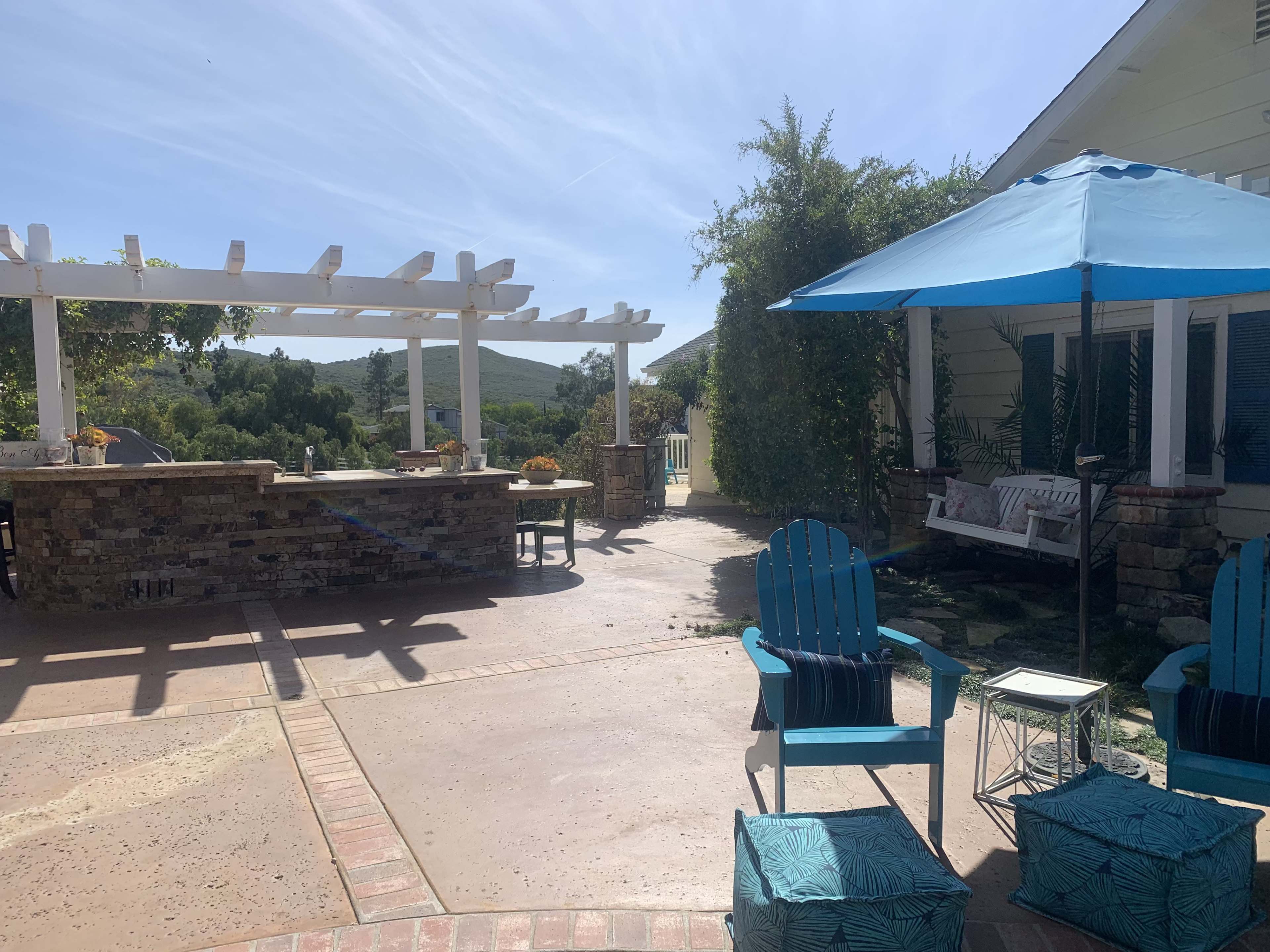 A patio area with a stone bar, blue chairs, an umbrella, and a seating area surrounded by greenery.