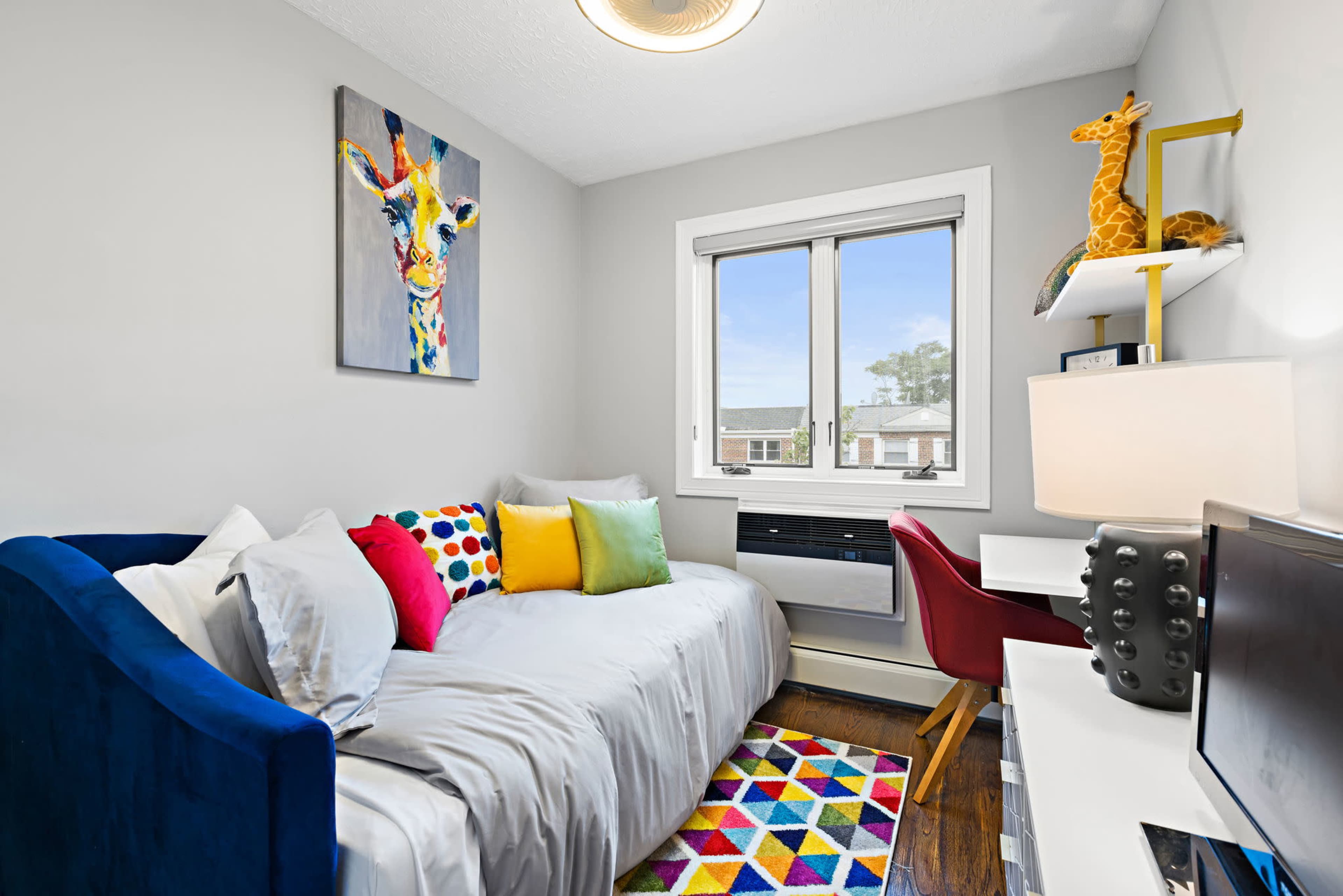 A brightly decorated small room features a daybed with colorful pillows, a patterned rug, a window with a view, and a desk setup.