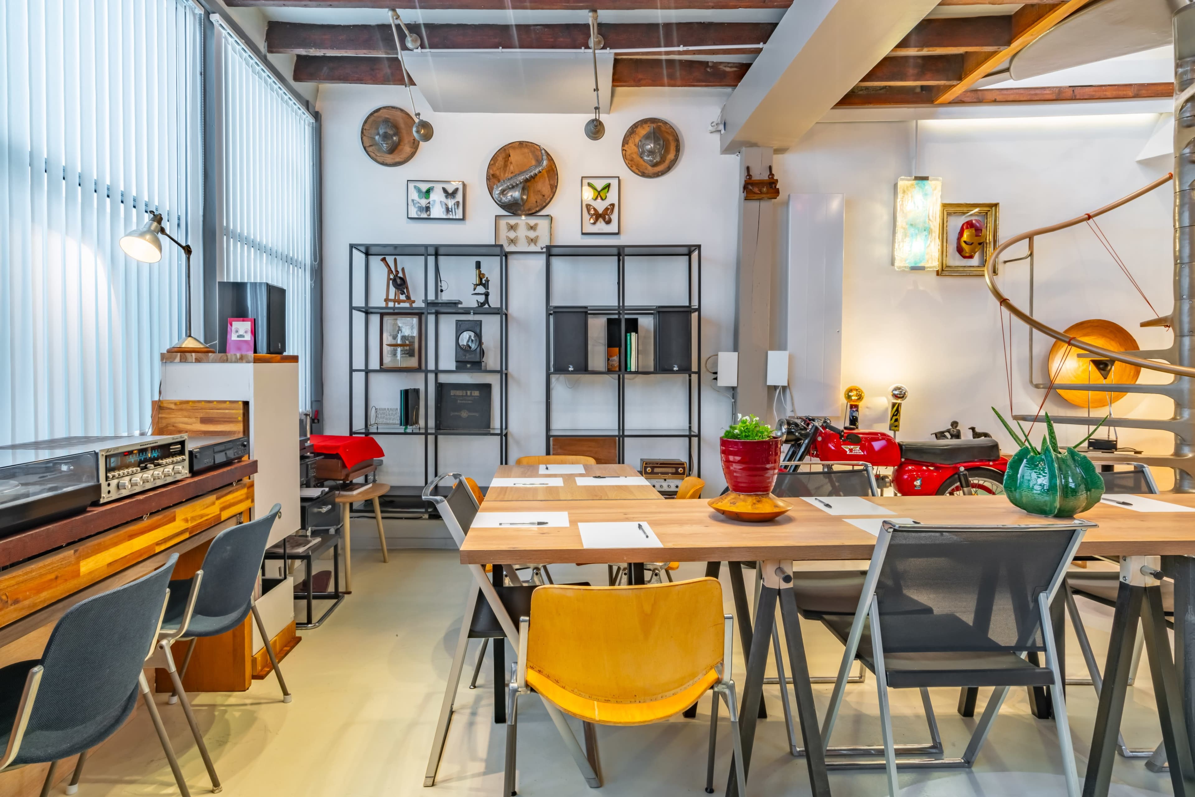 The image shows a modern, well-decorated workspace featuring wooden tables and chairs, shelves with various decorative items, and a spiral staircase.