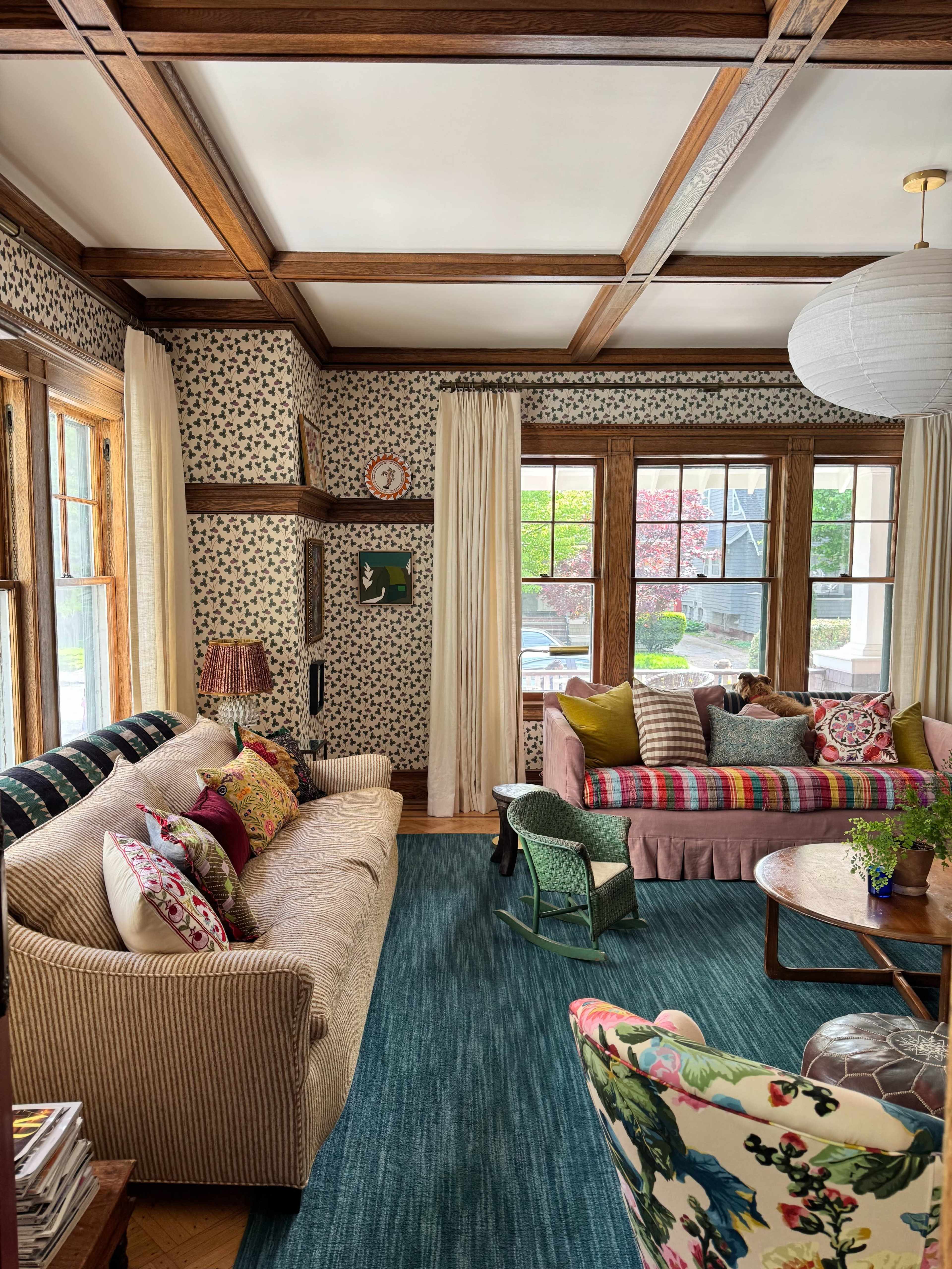 A cozy living room features patterned wallpaper, wooden beams, and two sofas adorned with colorful cushions, alongside a circular coffee table and plants.