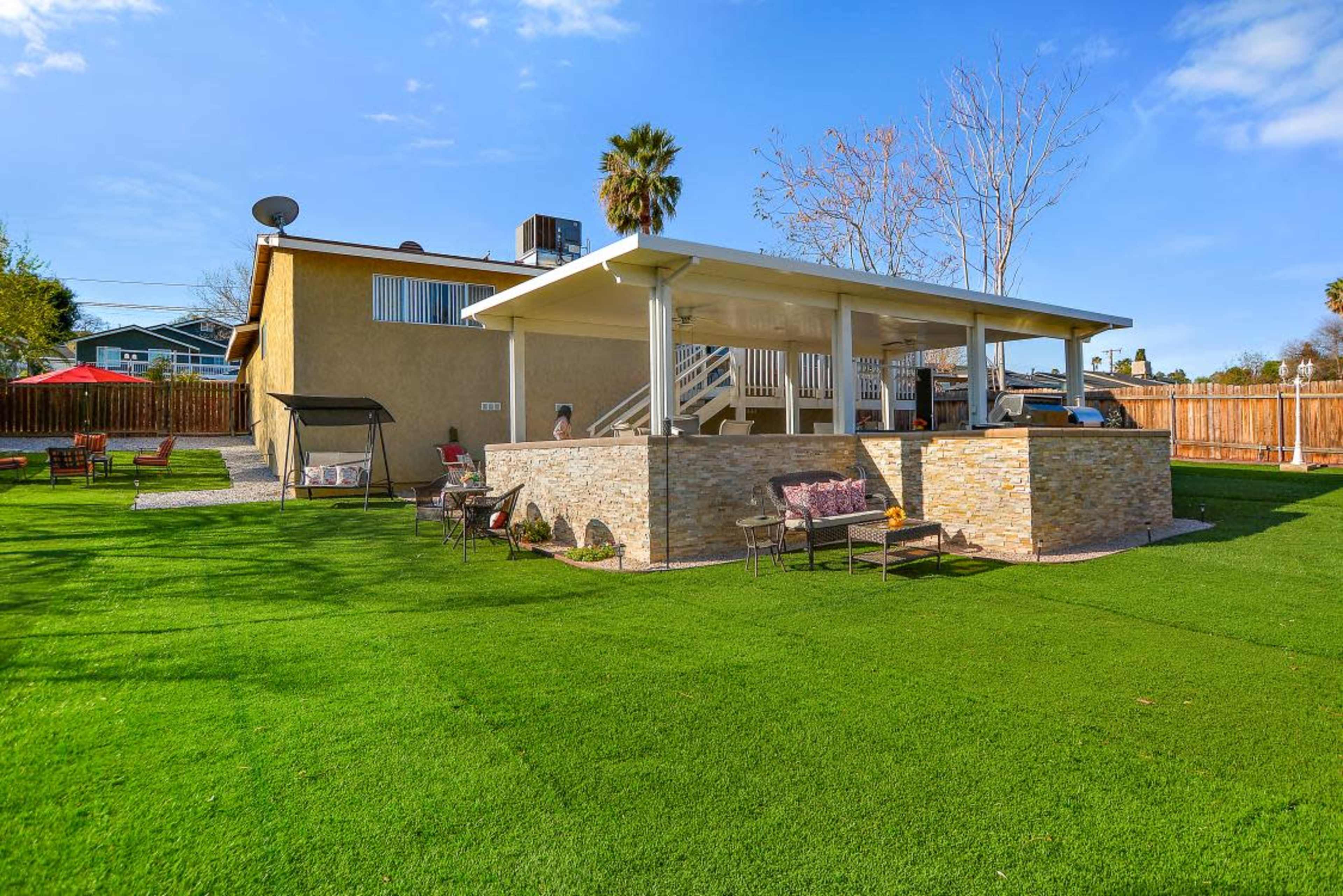 A backyard features a large, raised patio with stone walls, surrounded by green artificial grass and seating areas.
