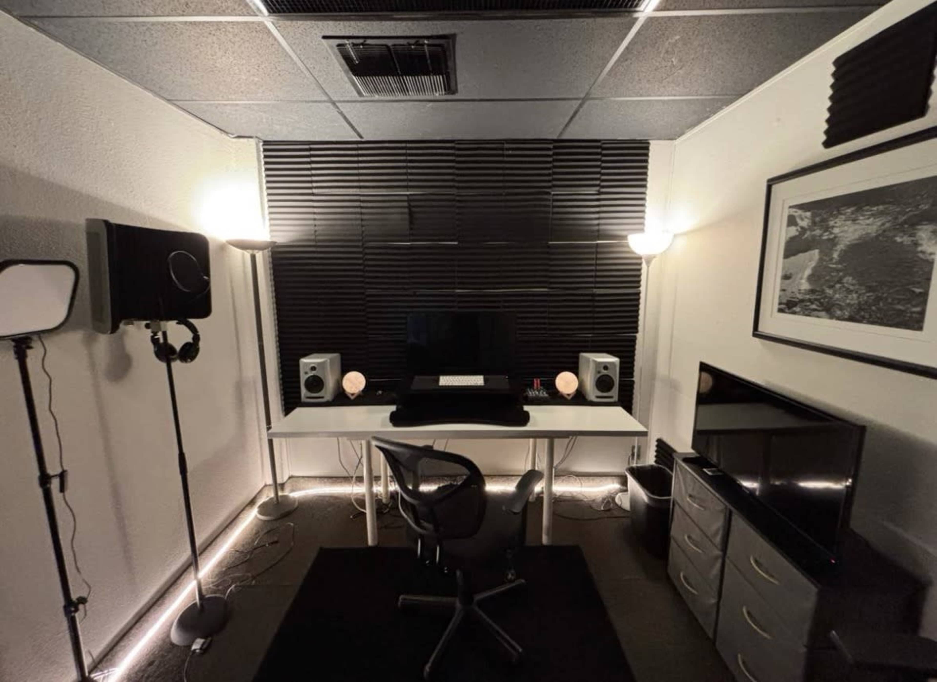 The image shows a small recording studio with soundproofing panels, a desk with a computer and speakers, and various lighting fixtures.