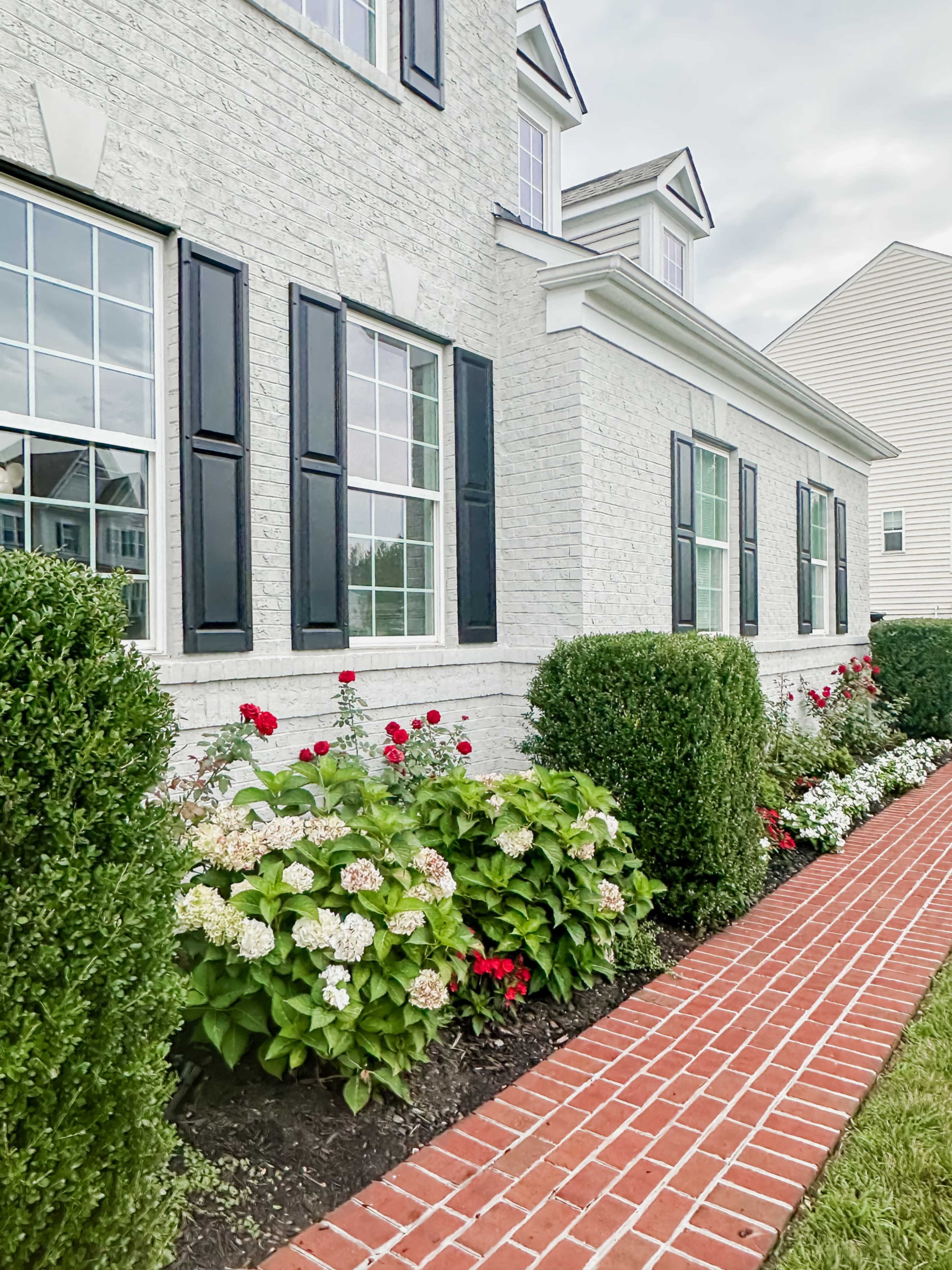 A brick walkway lined with neatly trimmed hedges and colorful flowers leads to the side of a white brick house with black shutters.