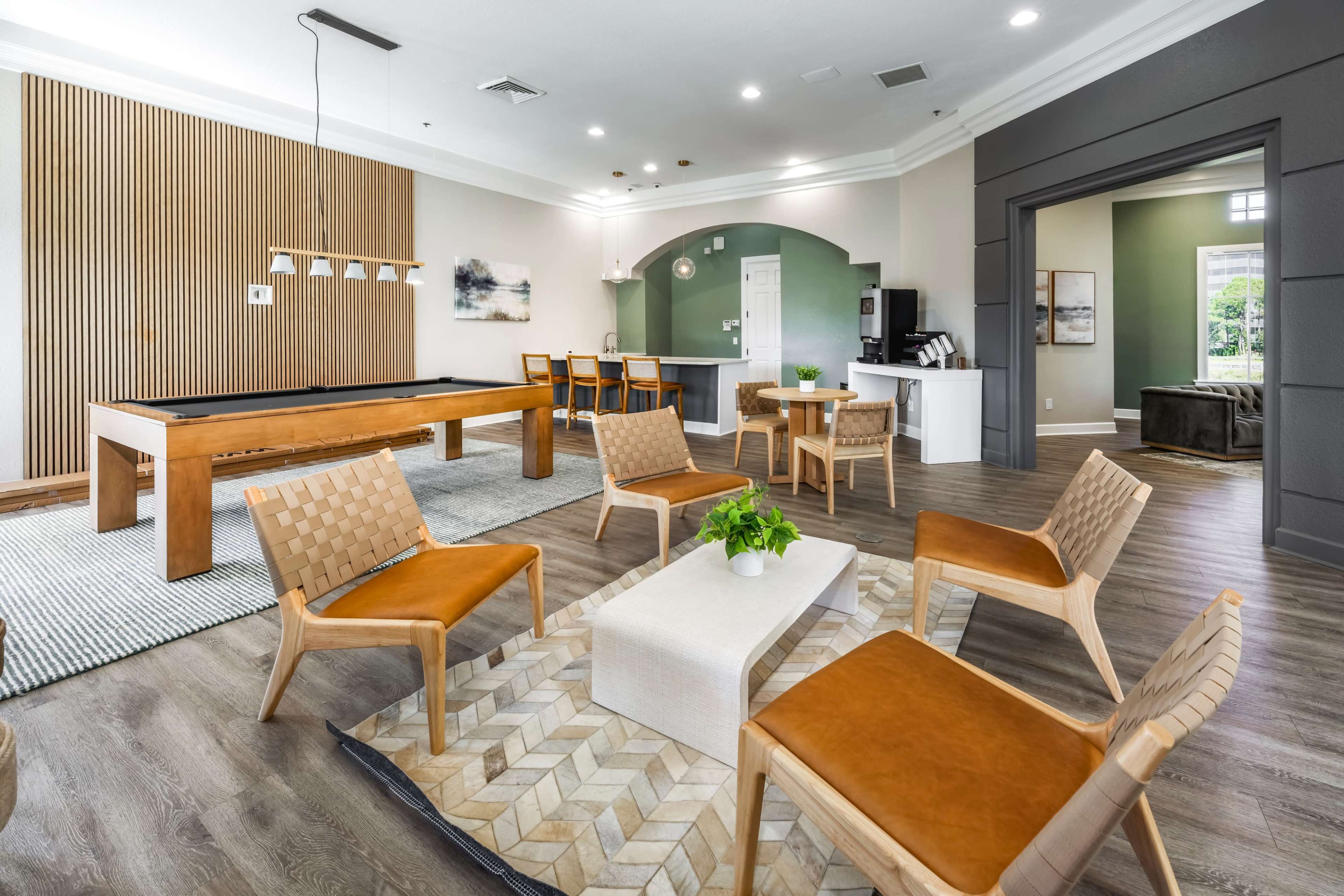 The image shows a modern lounge area featuring a pool table, several wooden chairs, a coffee table, and a kitchenette in a well-lit room.