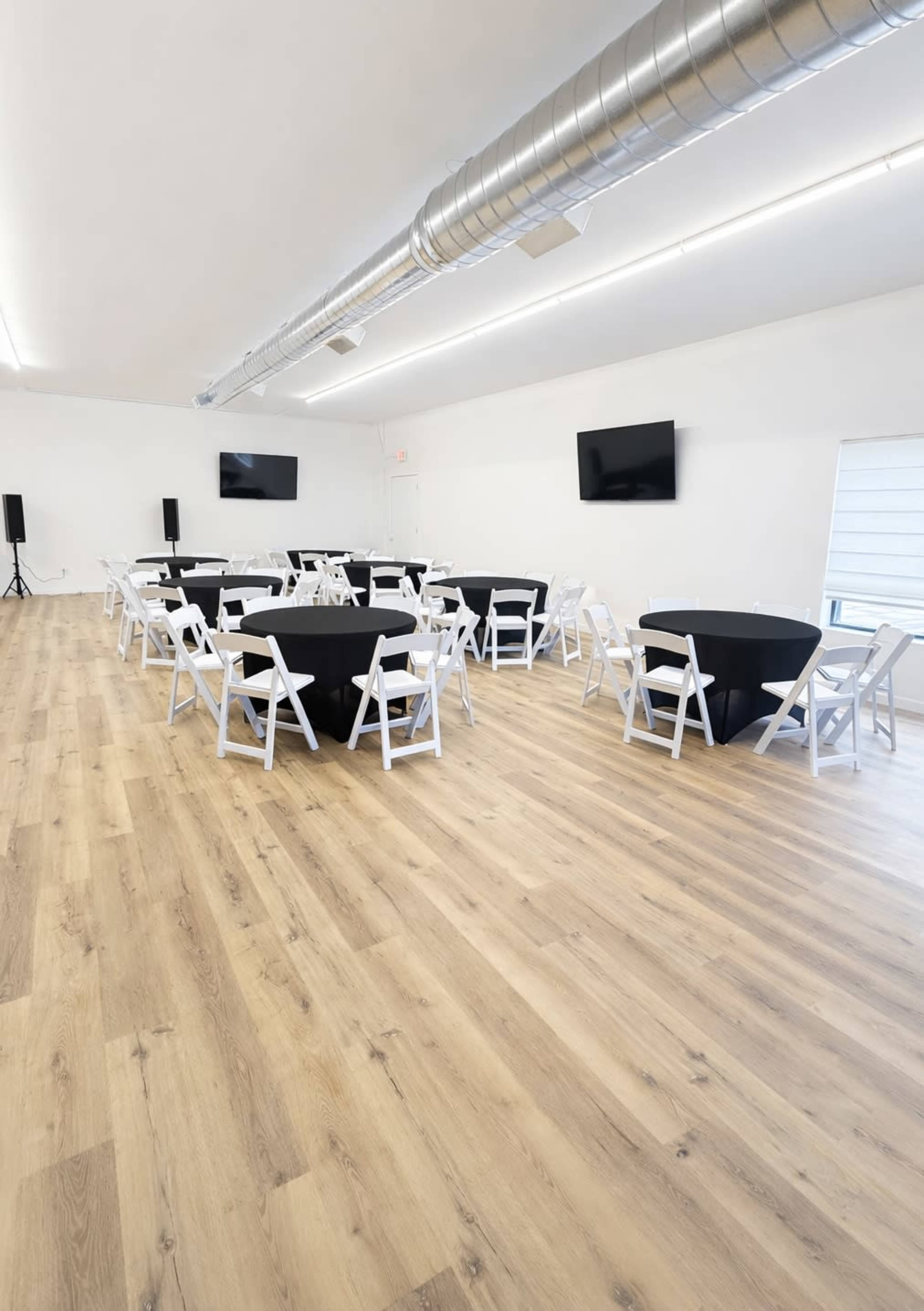 The image shows a spacious event room with several round black tables surrounded by white chairs, wooden flooring, and two wall-mounted televisions.