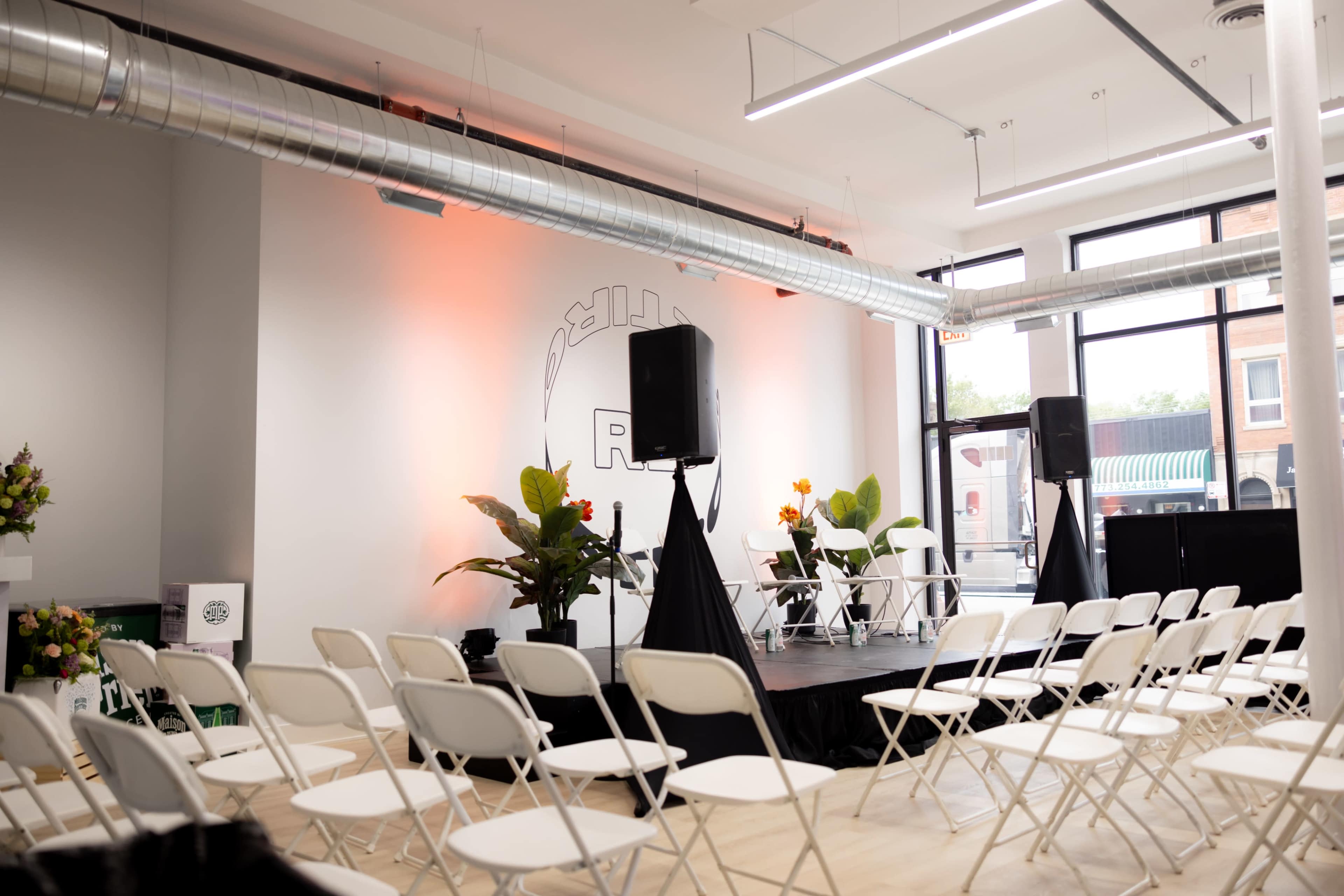 A stage with a black draped backdrop, flanked by plants and speakers, is set up in a modern event space with rows of white chairs facing it.