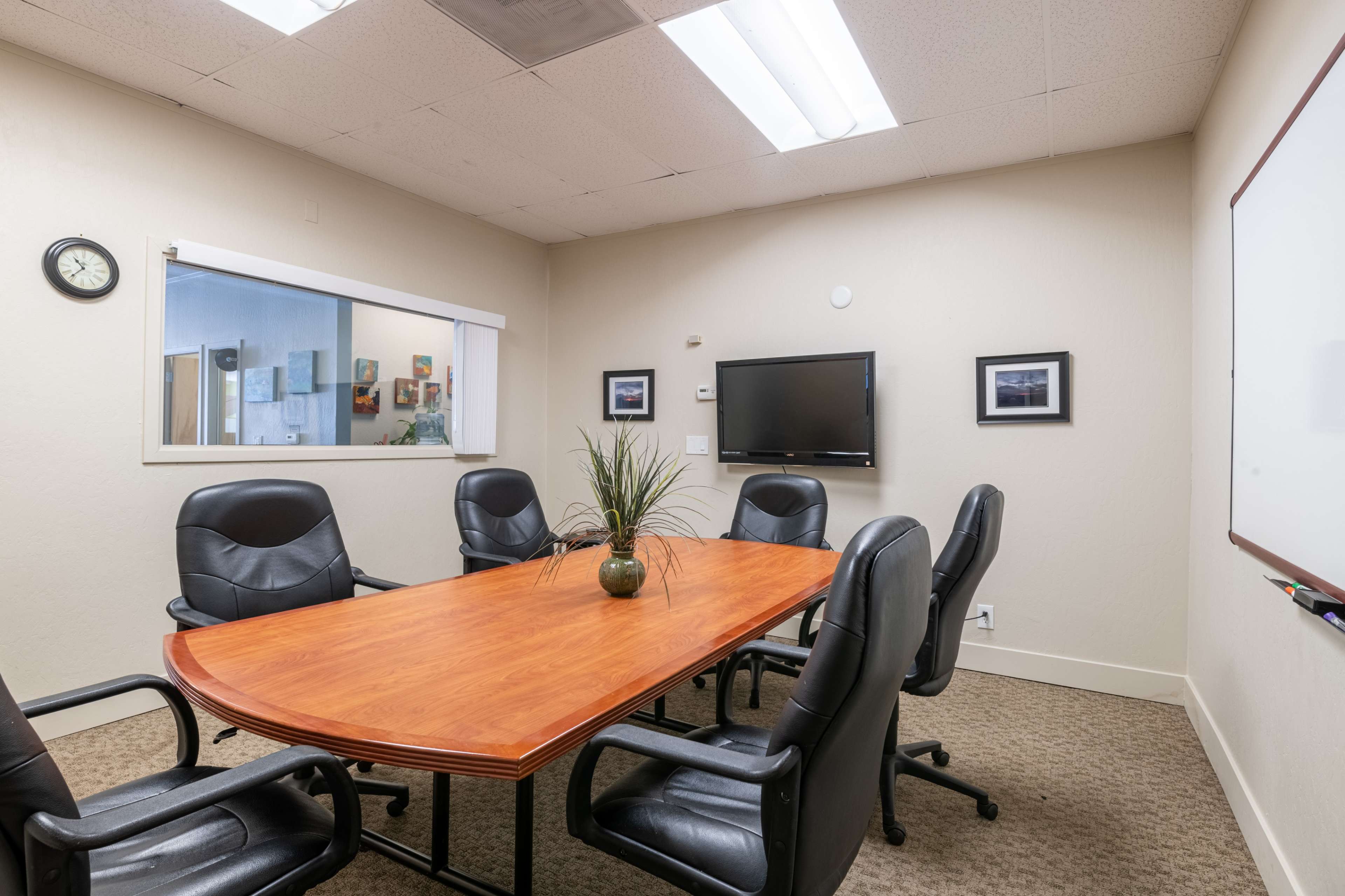 A conference room features a large wooden table surrounded by six black chairs, with a television and a plant centerpiece.