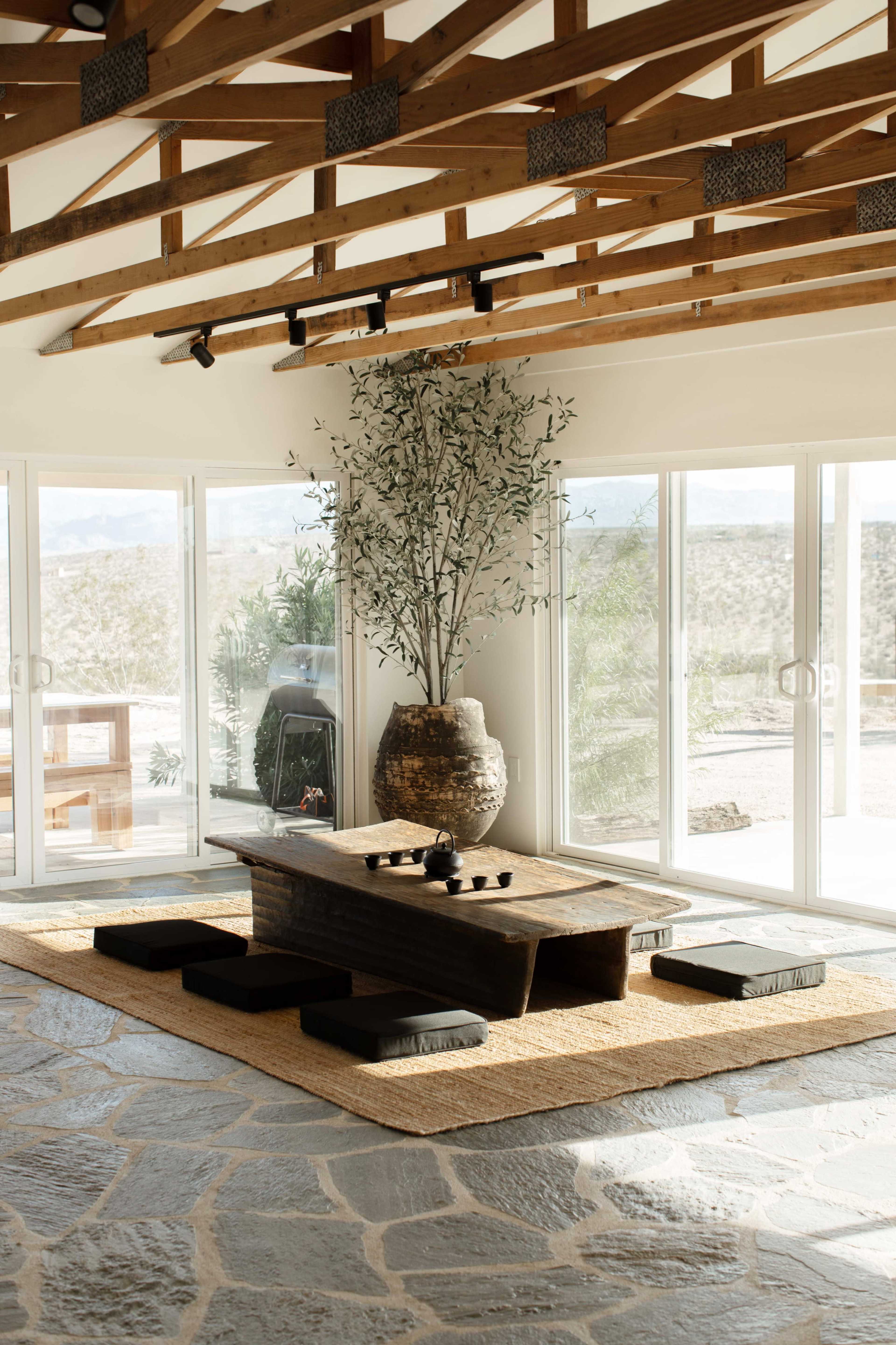 The interior features a stone floor, a large wooden table surrounded by floor cushions, and a tall plant in a decorative pot, with large glass doors allowing natural light and a view of the outdoors.