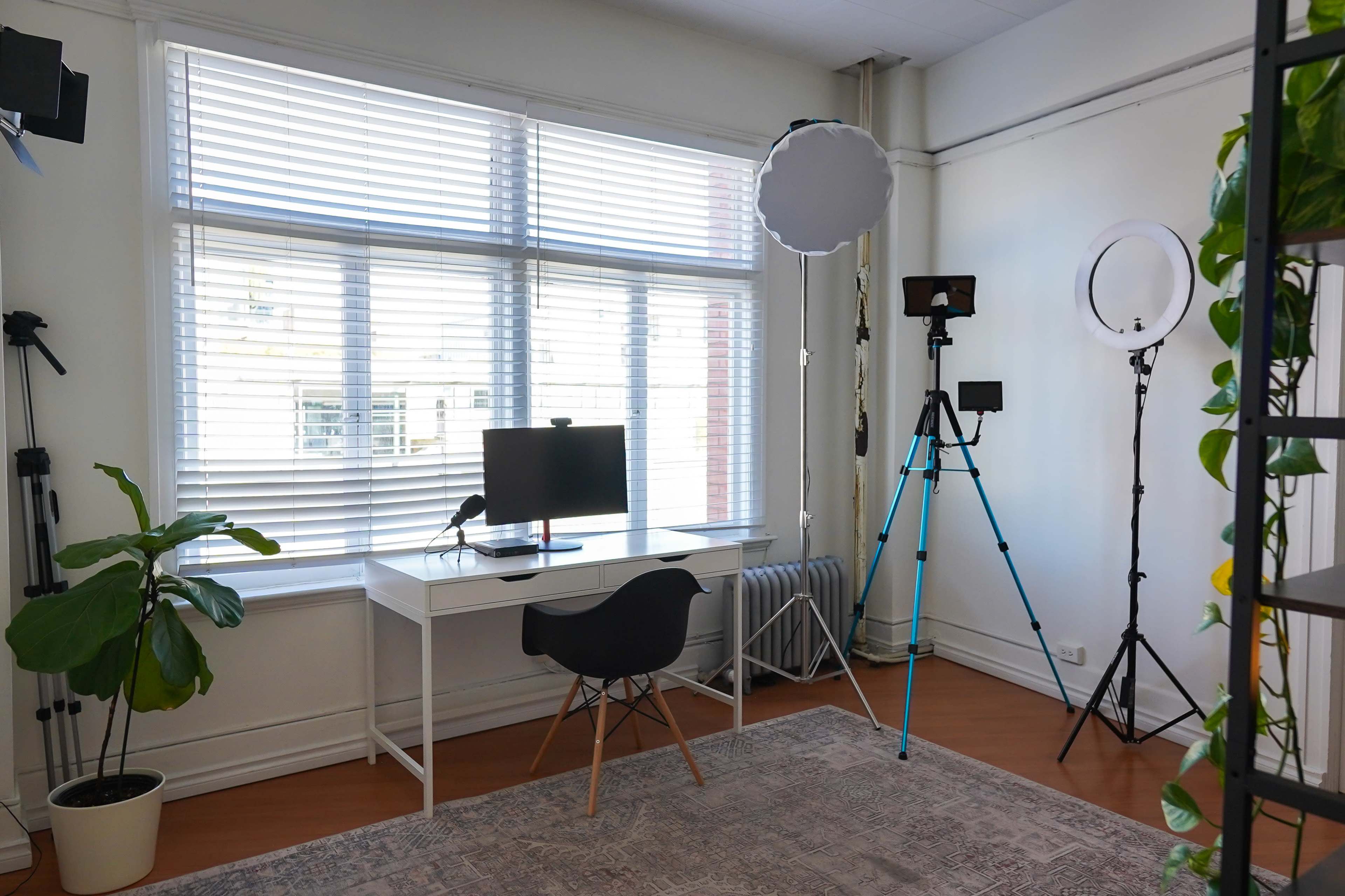 The room features a white desk with a computer, a black chair, and various lighting equipment beside a large window with blinds.