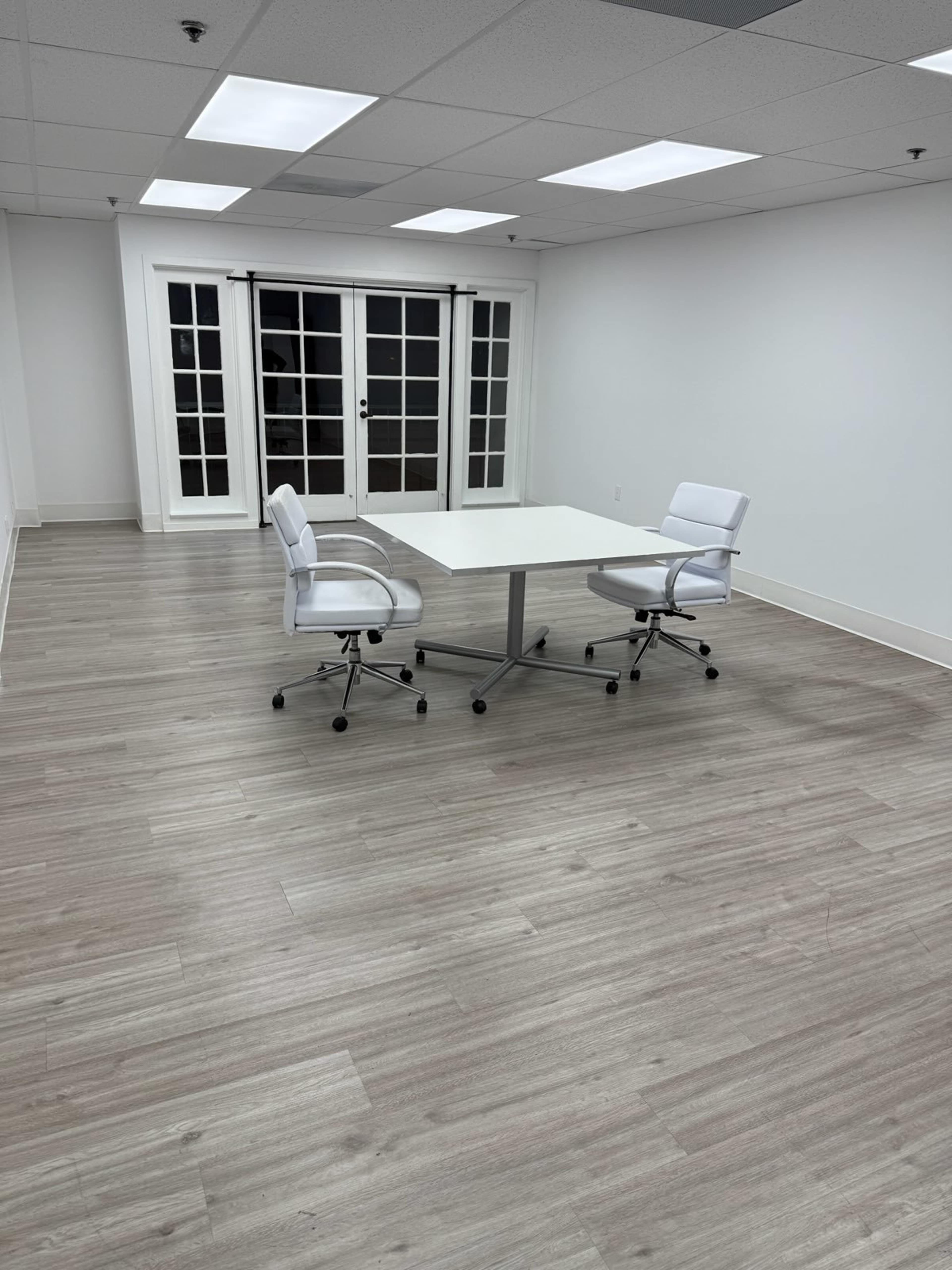 The image shows a minimalist office space with a square table and two white chairs, illuminated by overhead lighting and featuring large windows with French doors.