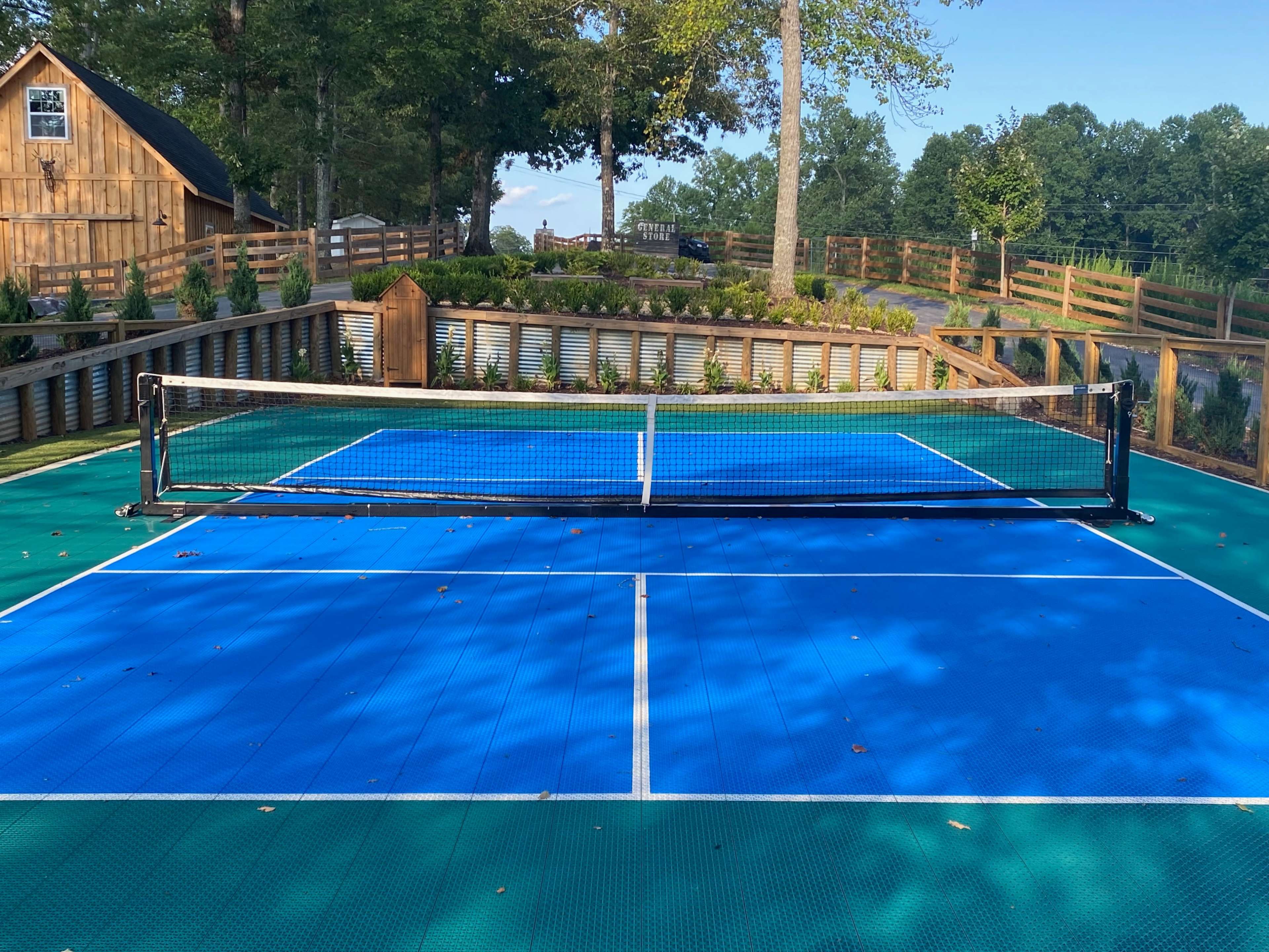 A tennis court with a blue surface and a net, surrounded by trees and a wooden fence.